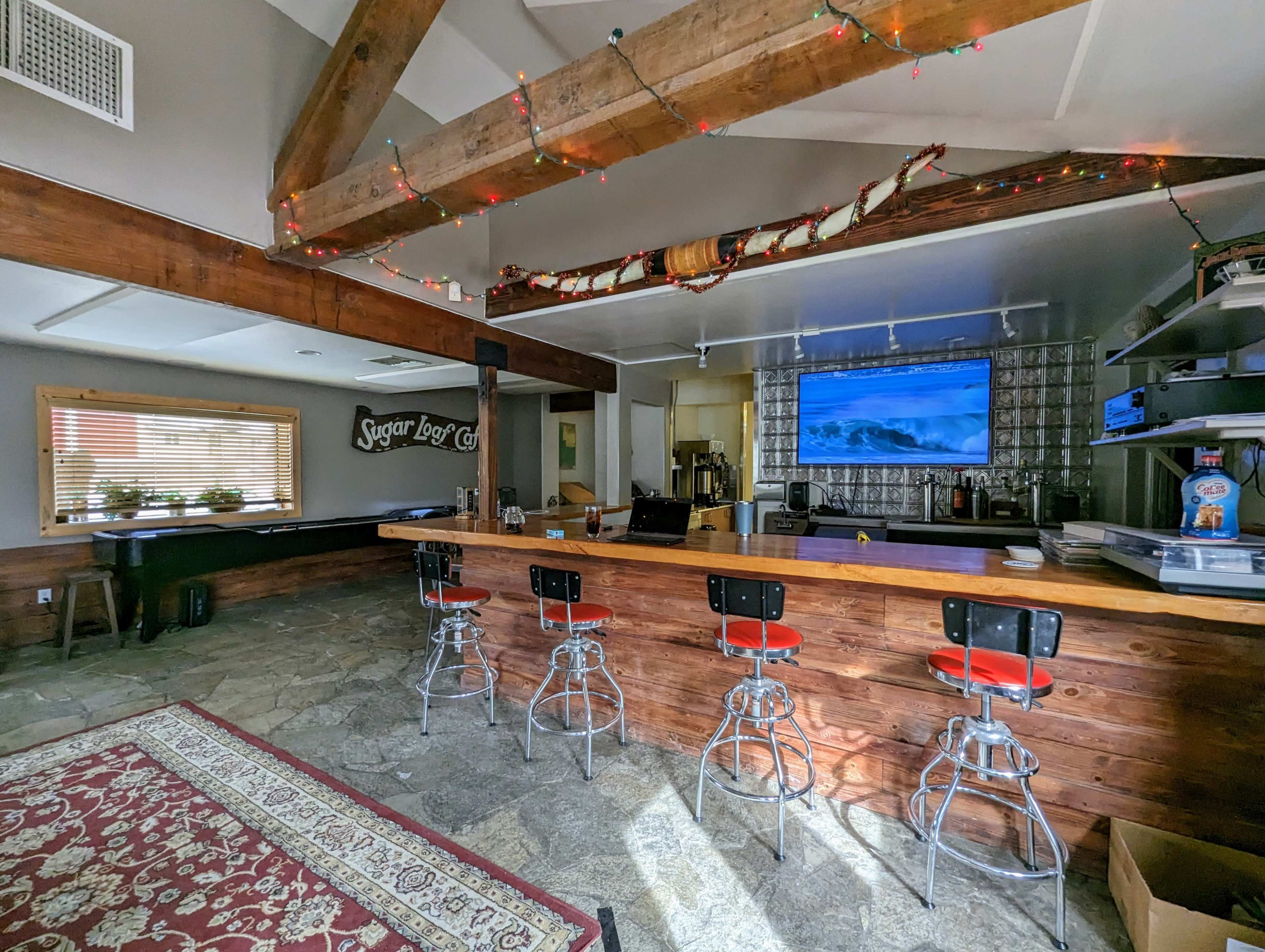 A rustic bar area with a stone floor, wooden beams, and high-backed stools in front of a long counter, featuring a large television displaying waves.