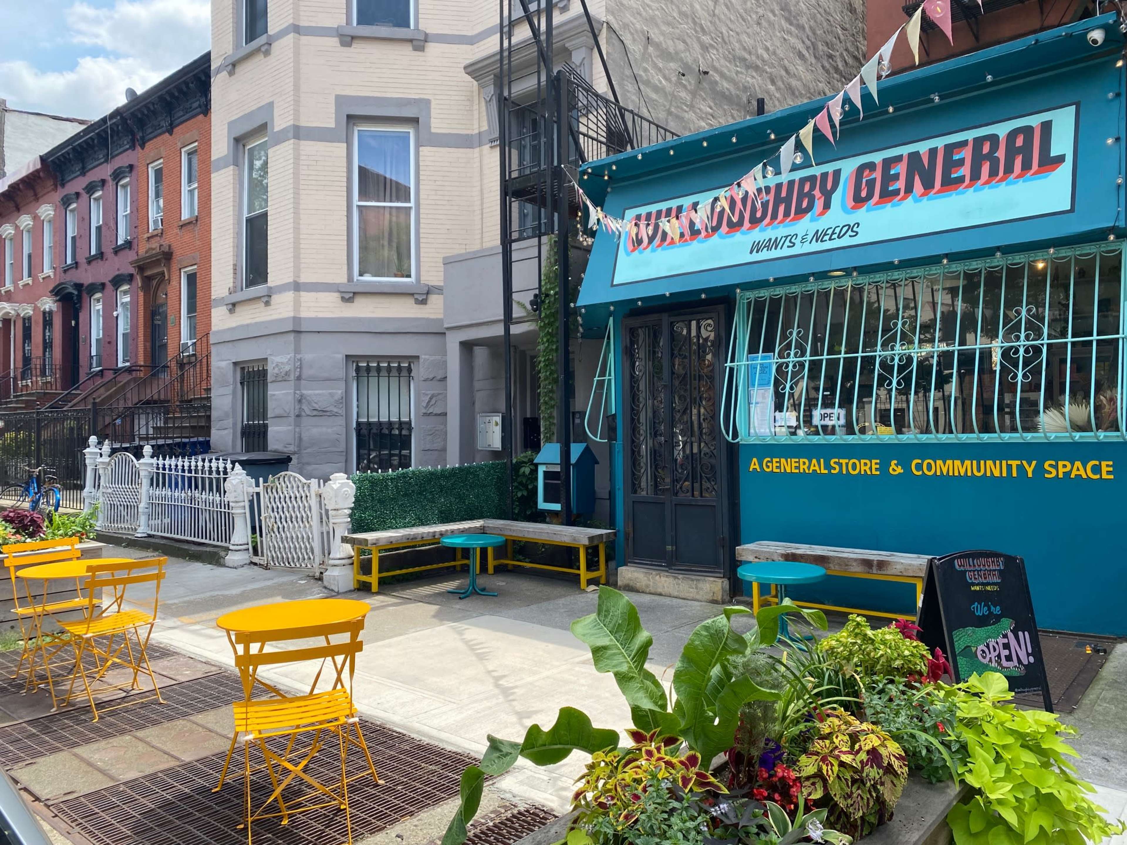 A colorful general store named "Gunningby General" next to brownstone buildings, featuring outdoor seating with yellow chairs on a sidewalk.