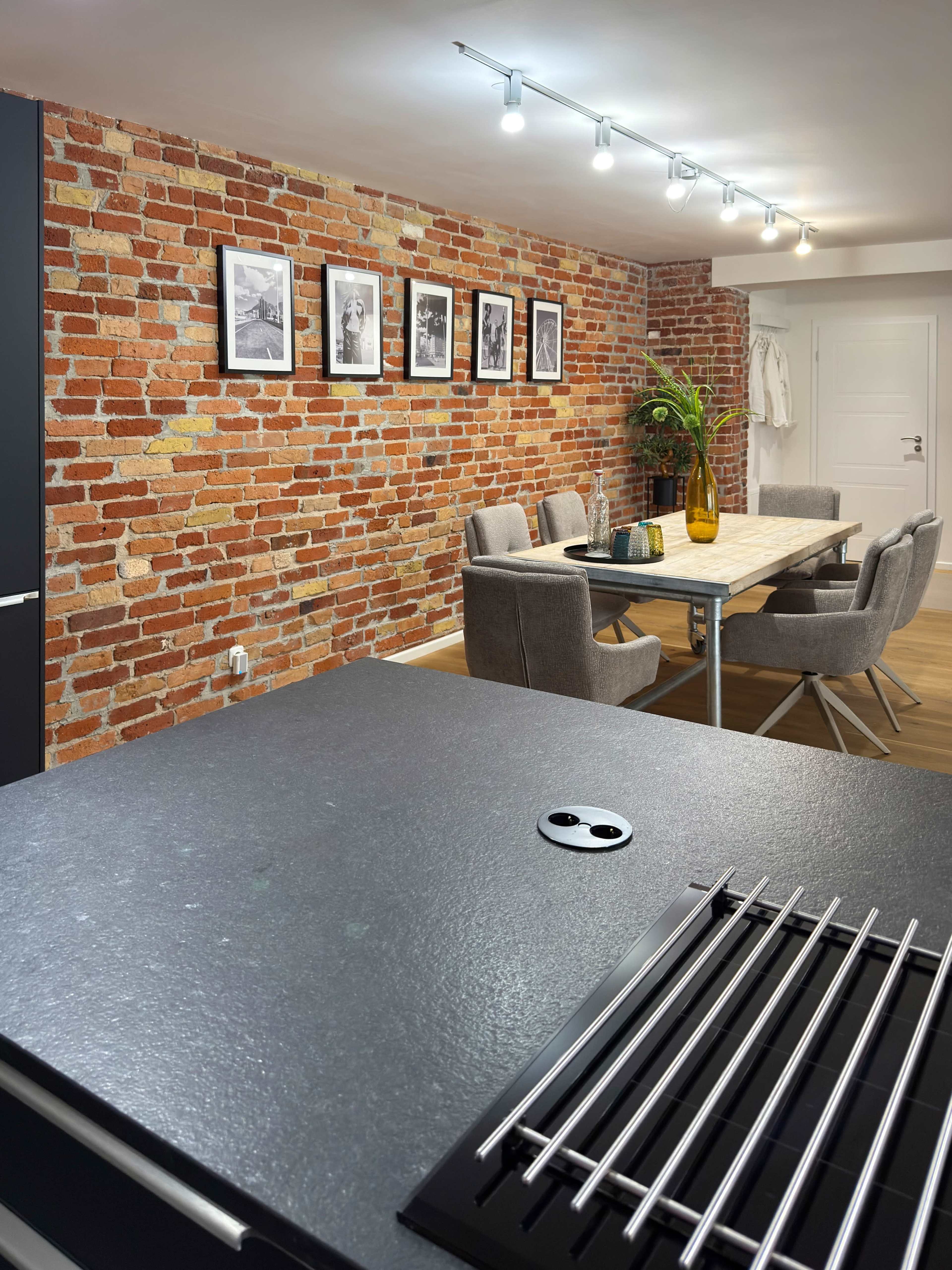 The image shows a modern kitchen with a dark countertop overlooking a dining area featuring a wooden table surrounded by gray chairs, and a brick wall adorned with black-and-white photographs.