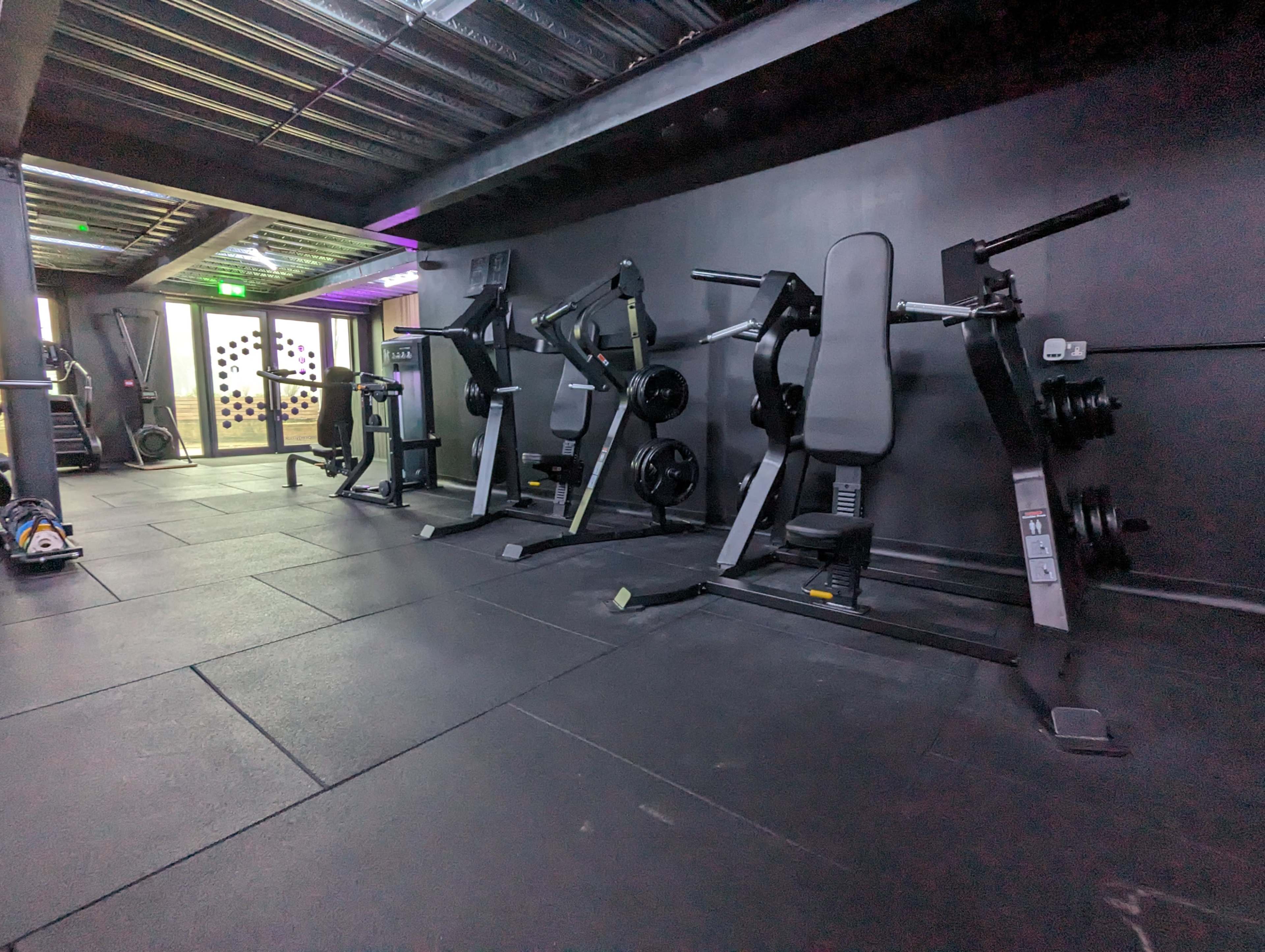 A gym interior with several fitness machines lined up against a black wall and a doorway leading to another space at the back.