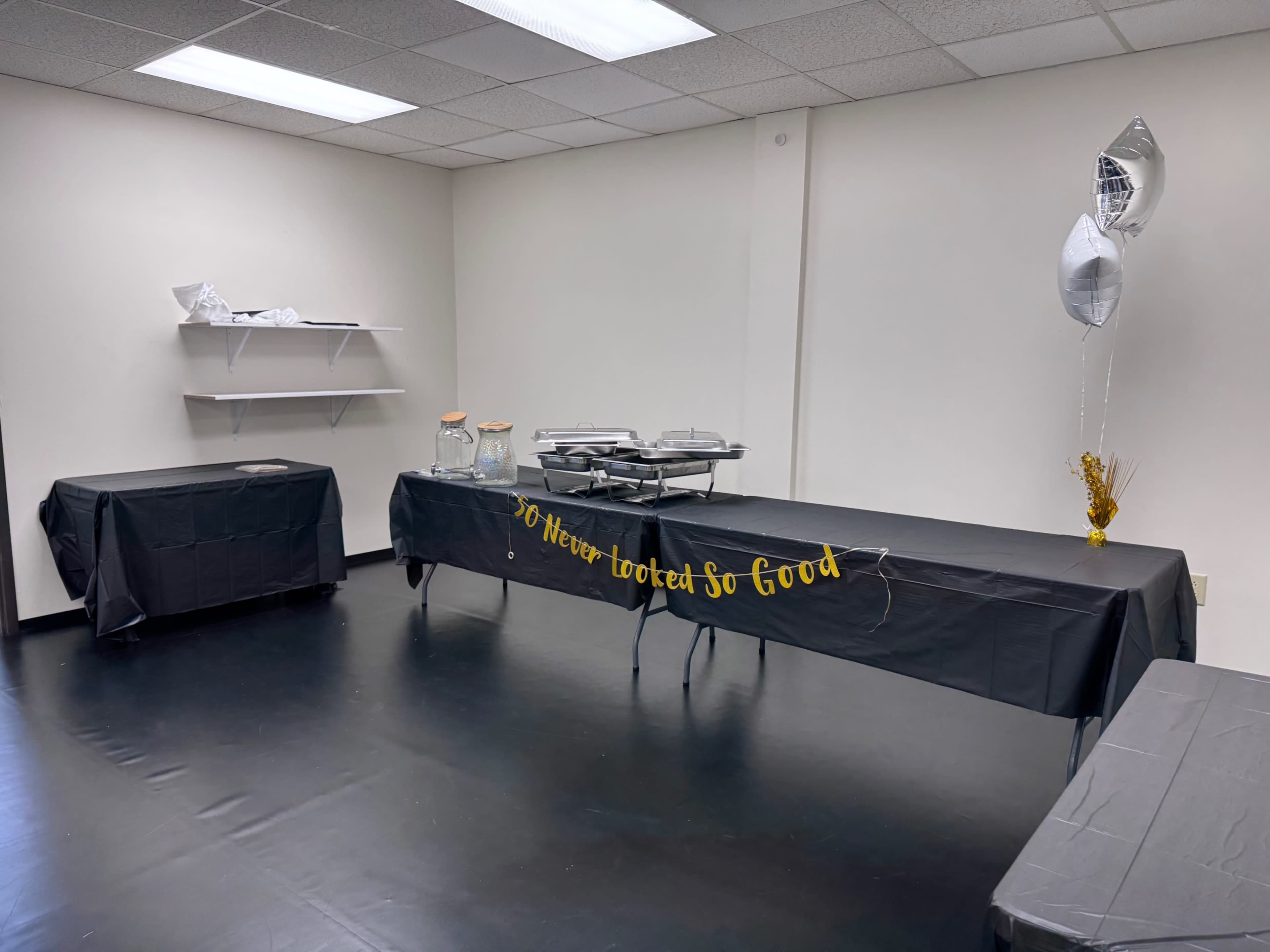 The image shows a partially decorated room with two black tables, one covered with food trays, and a banner that reads "You Never Tasted So Good."
