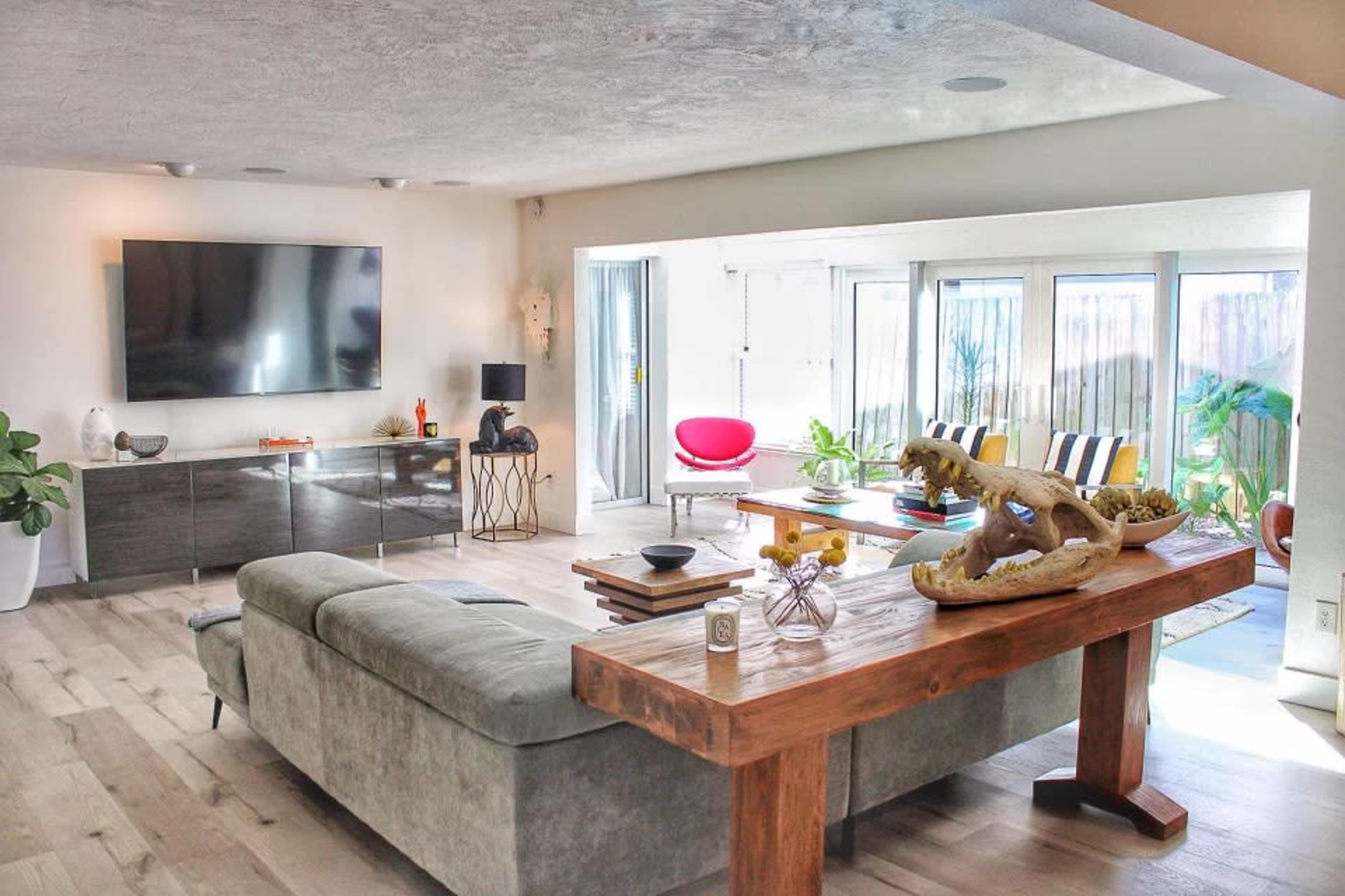 A modern living room with a large television mounted on the wall, a wooden dining table, a gray sofa, and large windows allowing natural light.