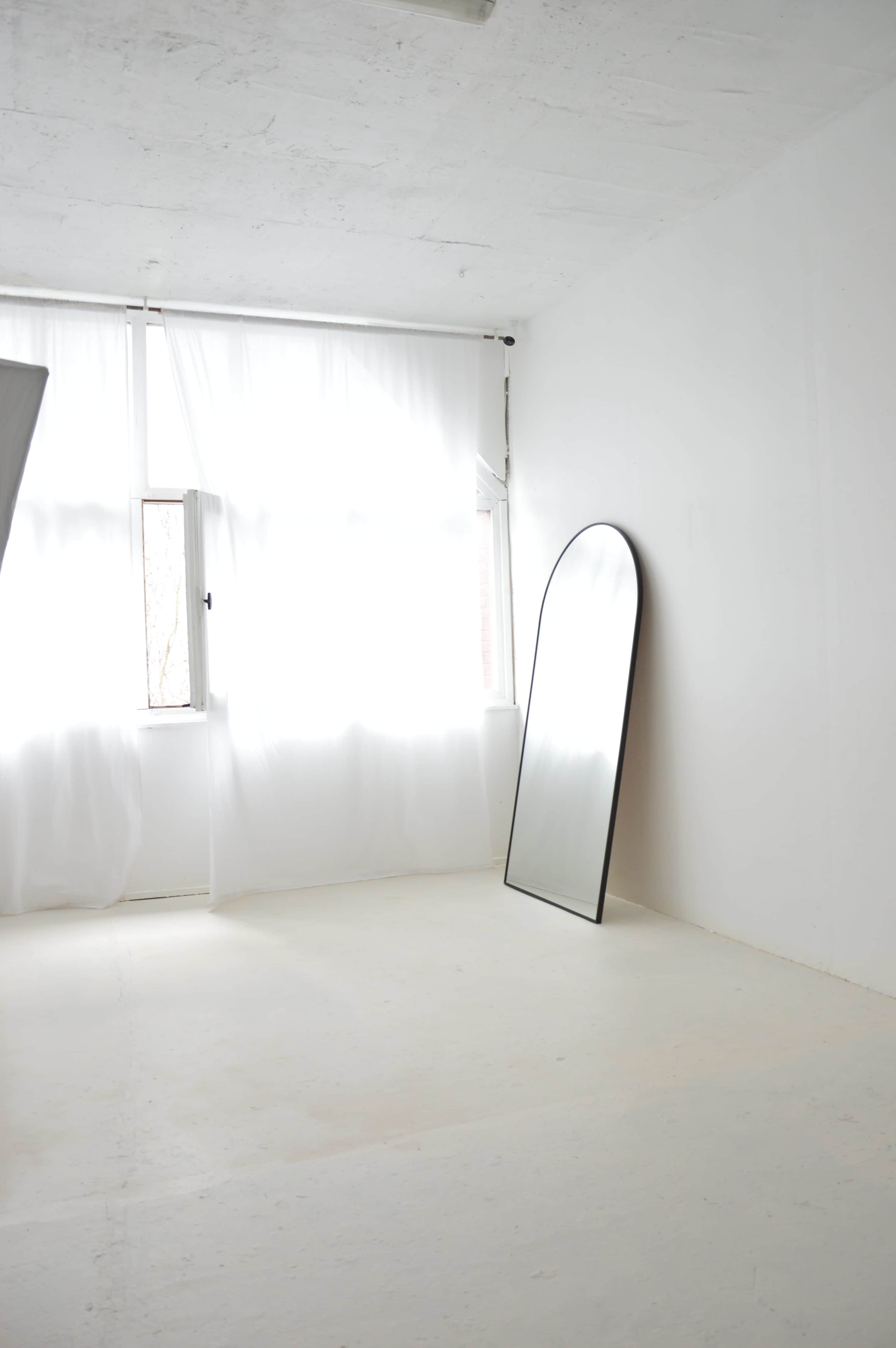 The image shows a minimalistic room with white walls, a large mirror leaning against the wall, and natural light coming through a window covered by sheer curtains.