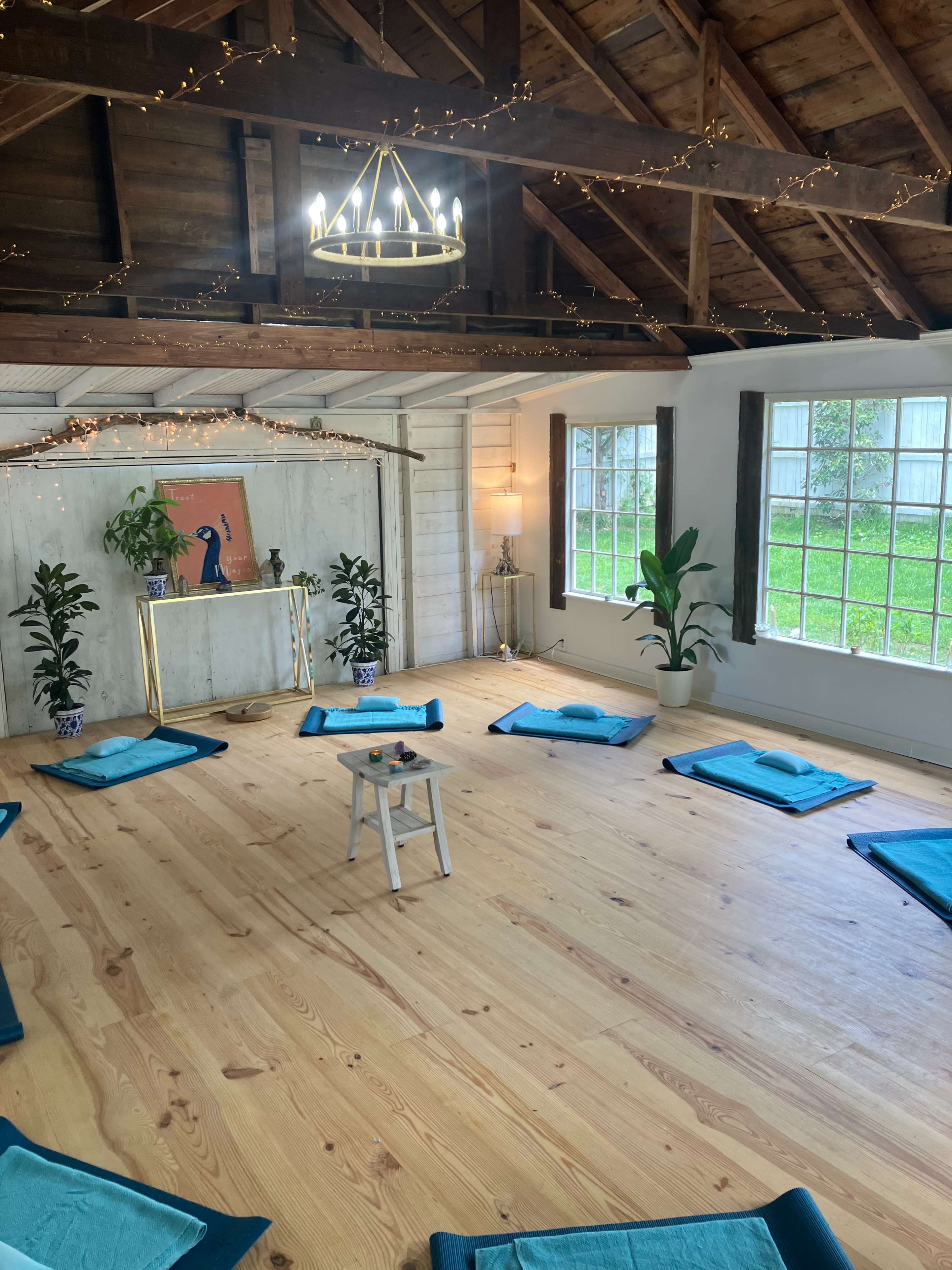 The image shows a serene yoga studio with blue mats arranged in a circular pattern around a small table, adorned with plants and decorative lighting.