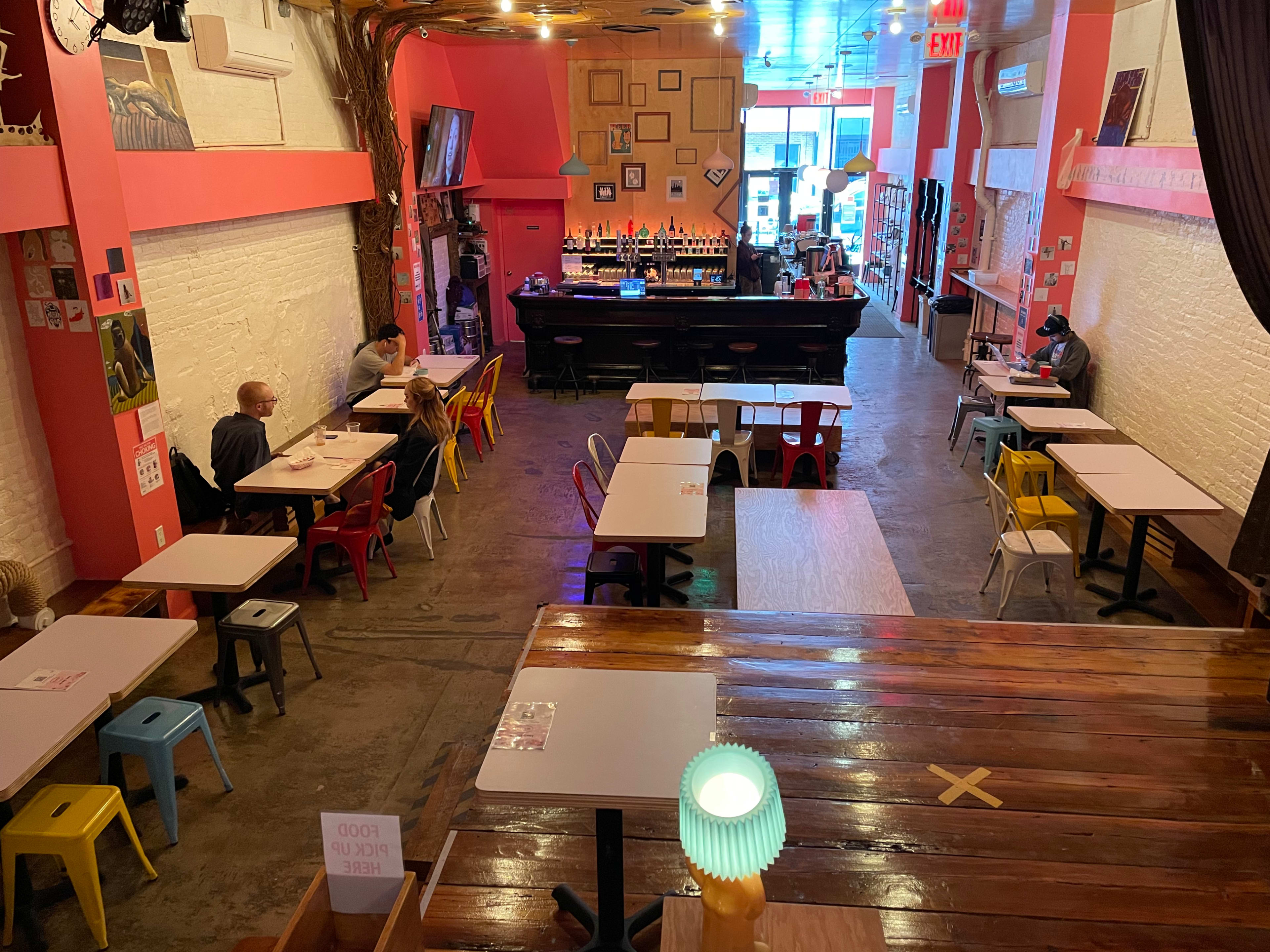 A brightly colored café interior with several small tables and chairs, a bar at the back, and patrons seated at the tables.