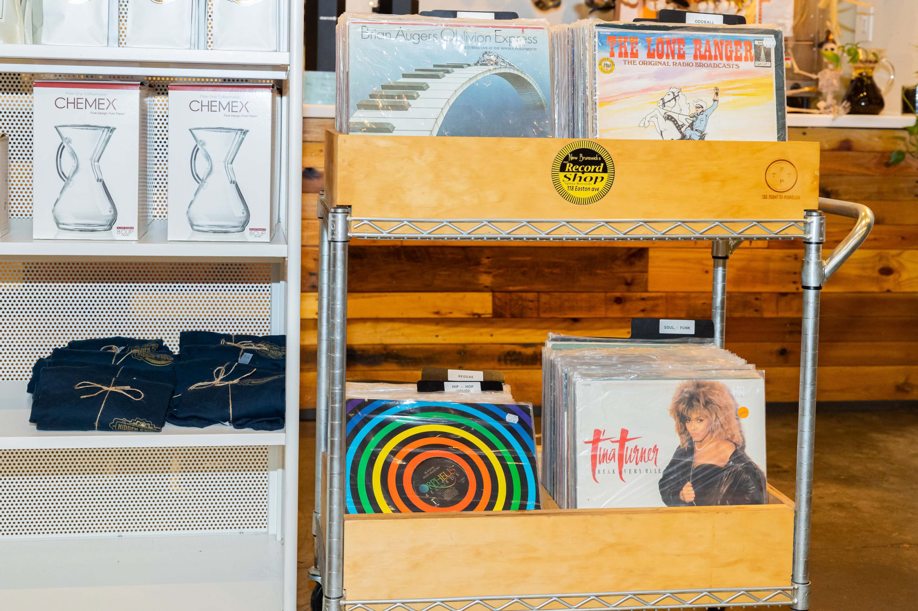 A wooden display cart holds various vinyl records beside a shelf of glass pitchers and folded clothing.