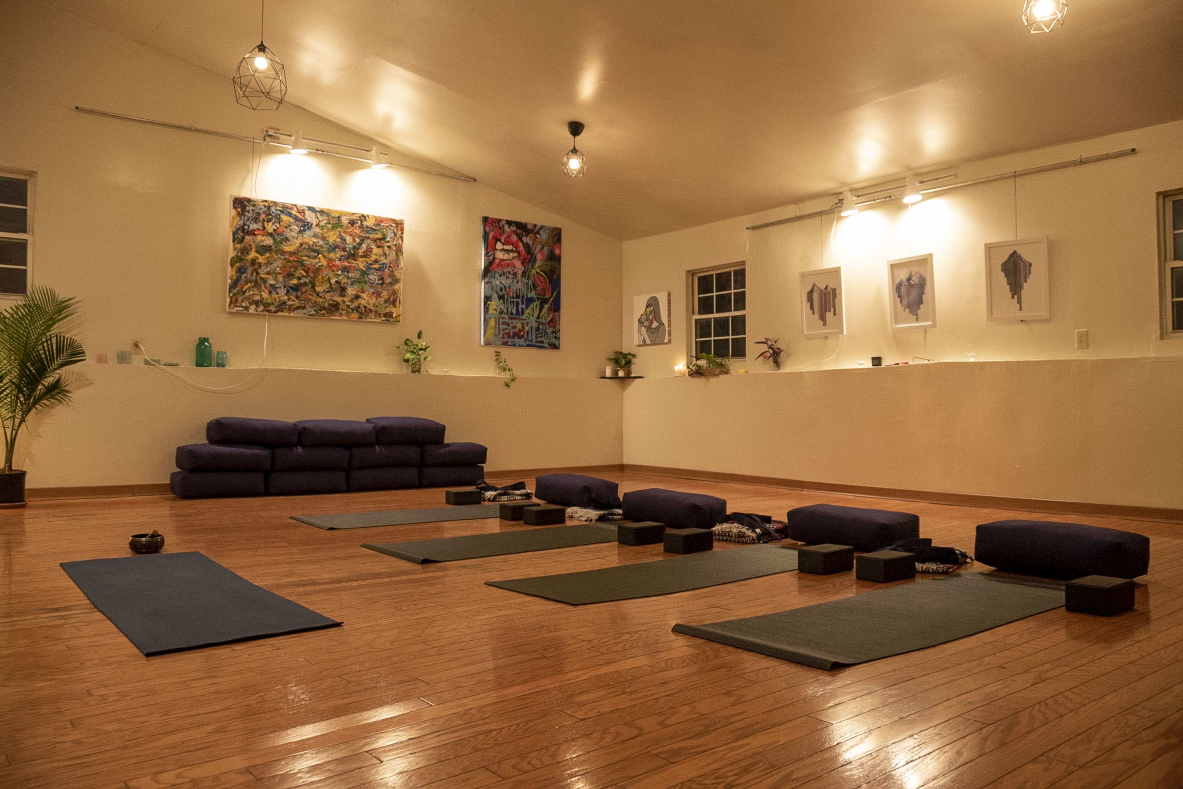 The image shows a spacious yoga studio with wooden floors, several yoga mats arranged in rows, and a couch along the back wall, adorned with artwork and plants.