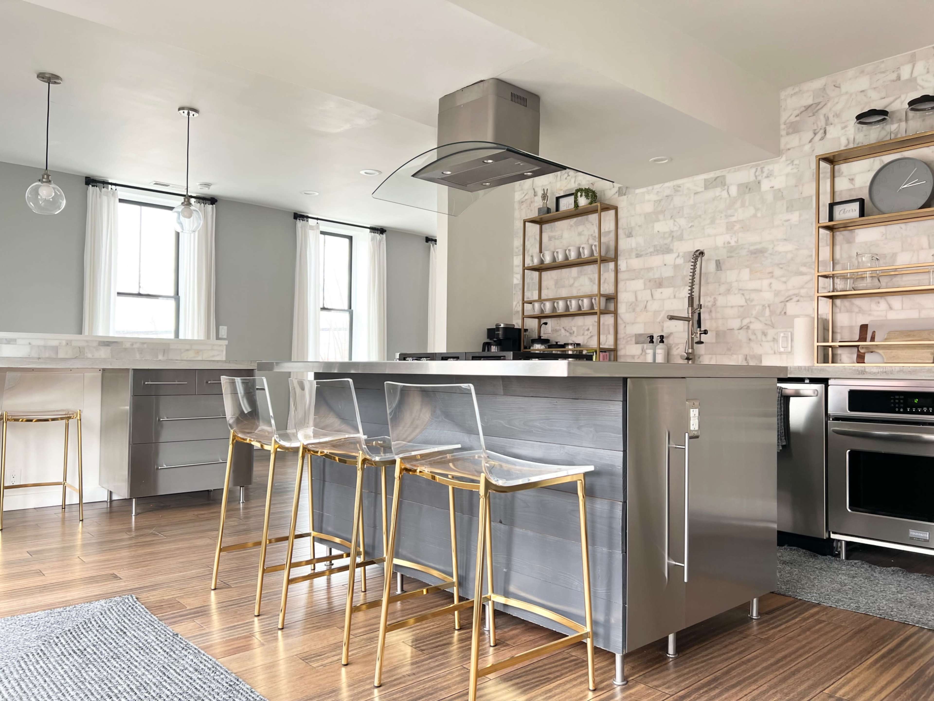 The kitchen features a modern stainless steel island with clear acrylic stools and a marble backsplash.