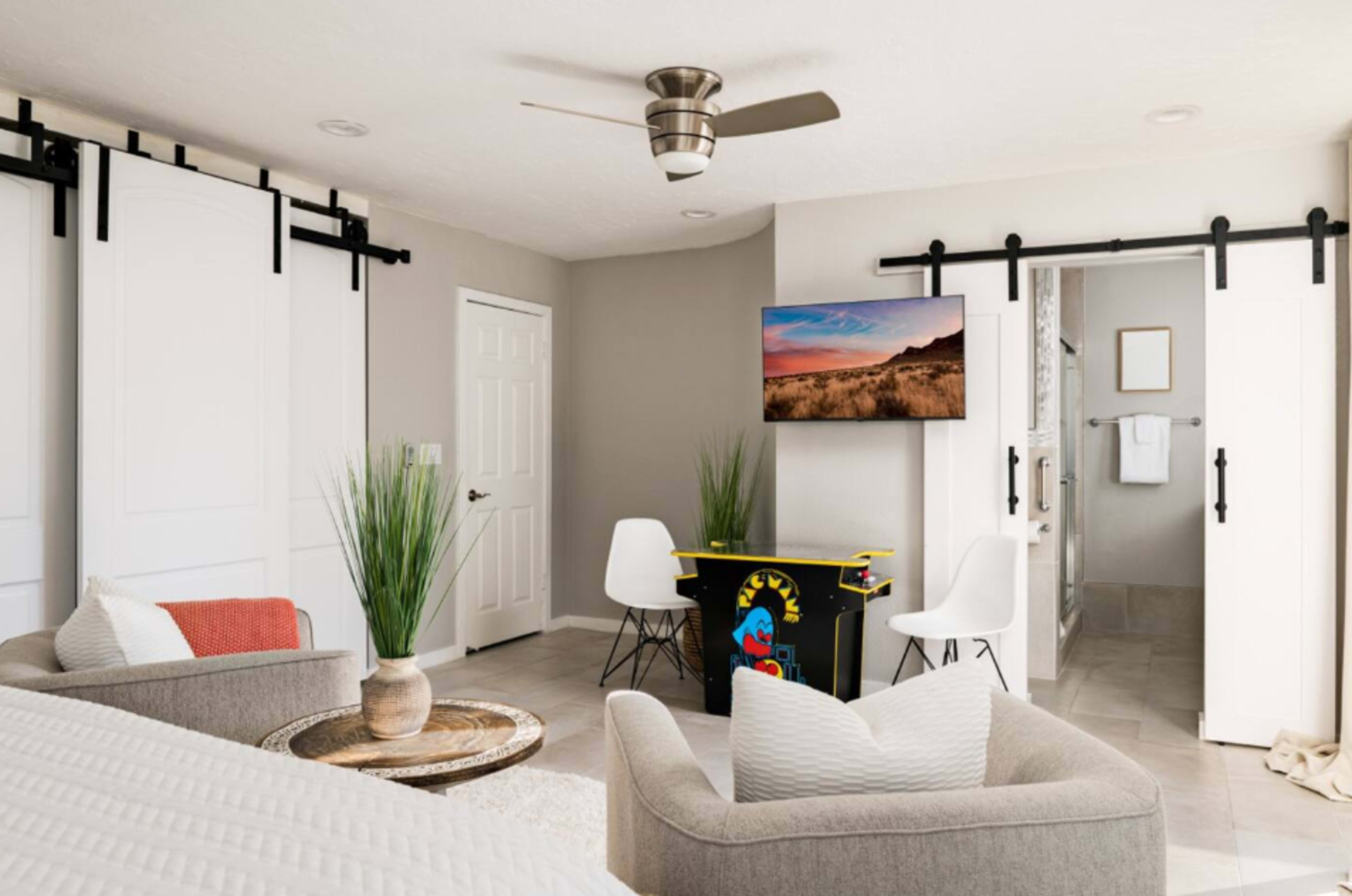 A cozy living space features a retro arcade game, a small seating area with two armchairs, and a television mounted on the wall, all surrounded by neutral-colored walls and decorative plants.