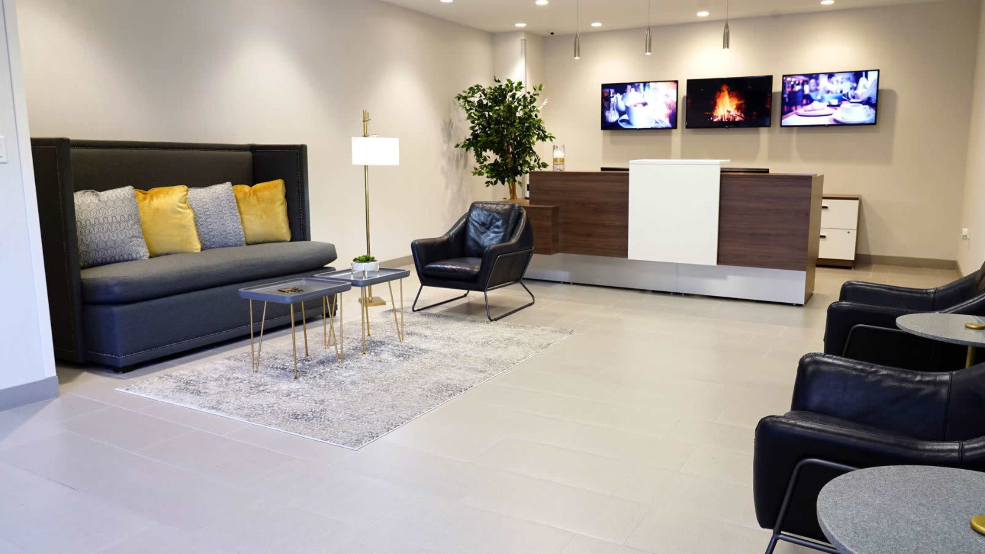 A modern waiting area featuring a dark sofa with yellow pillows, two black chairs, a coffee table, and a reception desk, with screens displaying content on the wall.