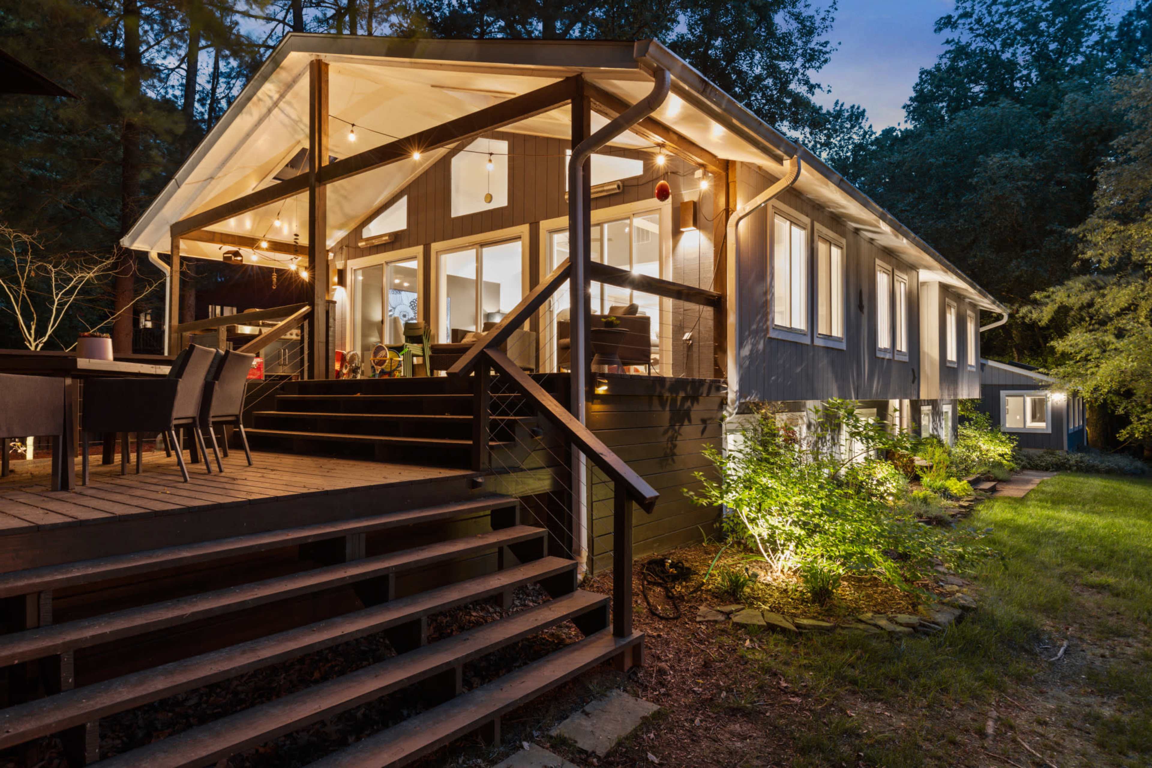 A modern house with large windows and a deck is illuminated at dusk, surrounded by trees.