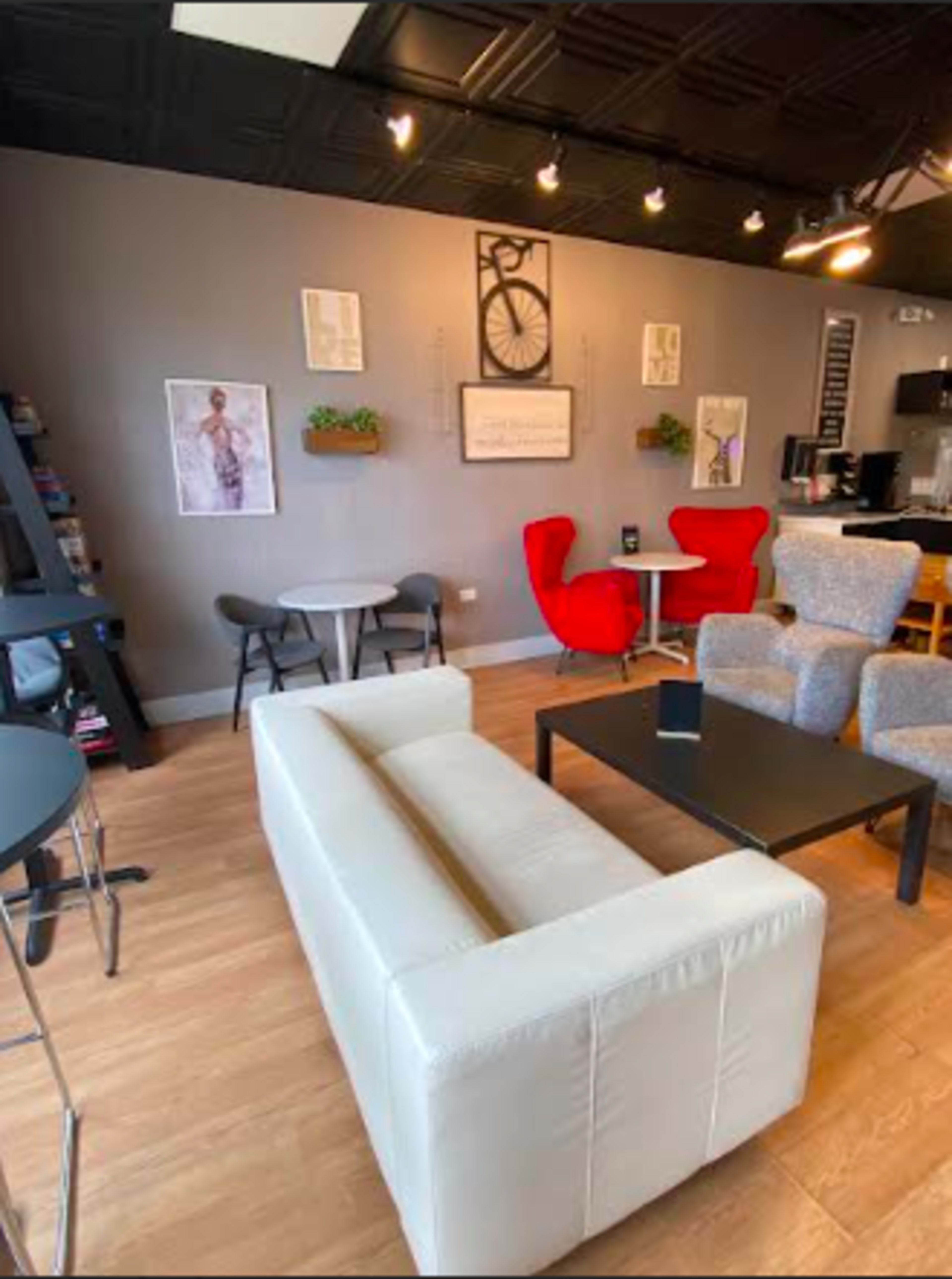 The image shows a modern café interior featuring a white sofa, a black coffee table, red and gray armchairs, and wall decor including framed art and plants.