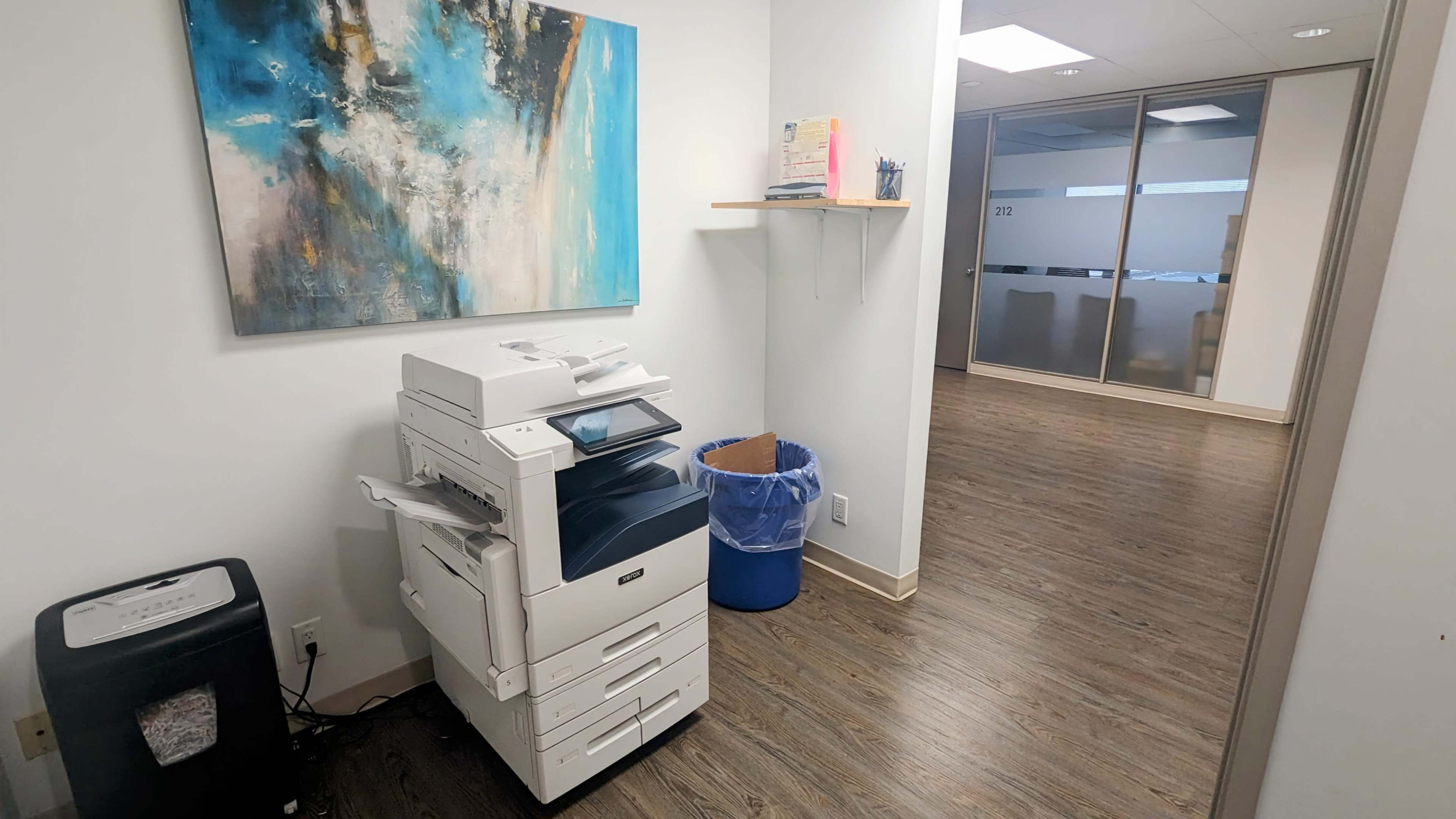 A photocopier stands next to a blue trash bin in a brightly lit office corridor with a large abstract painting on the wall.