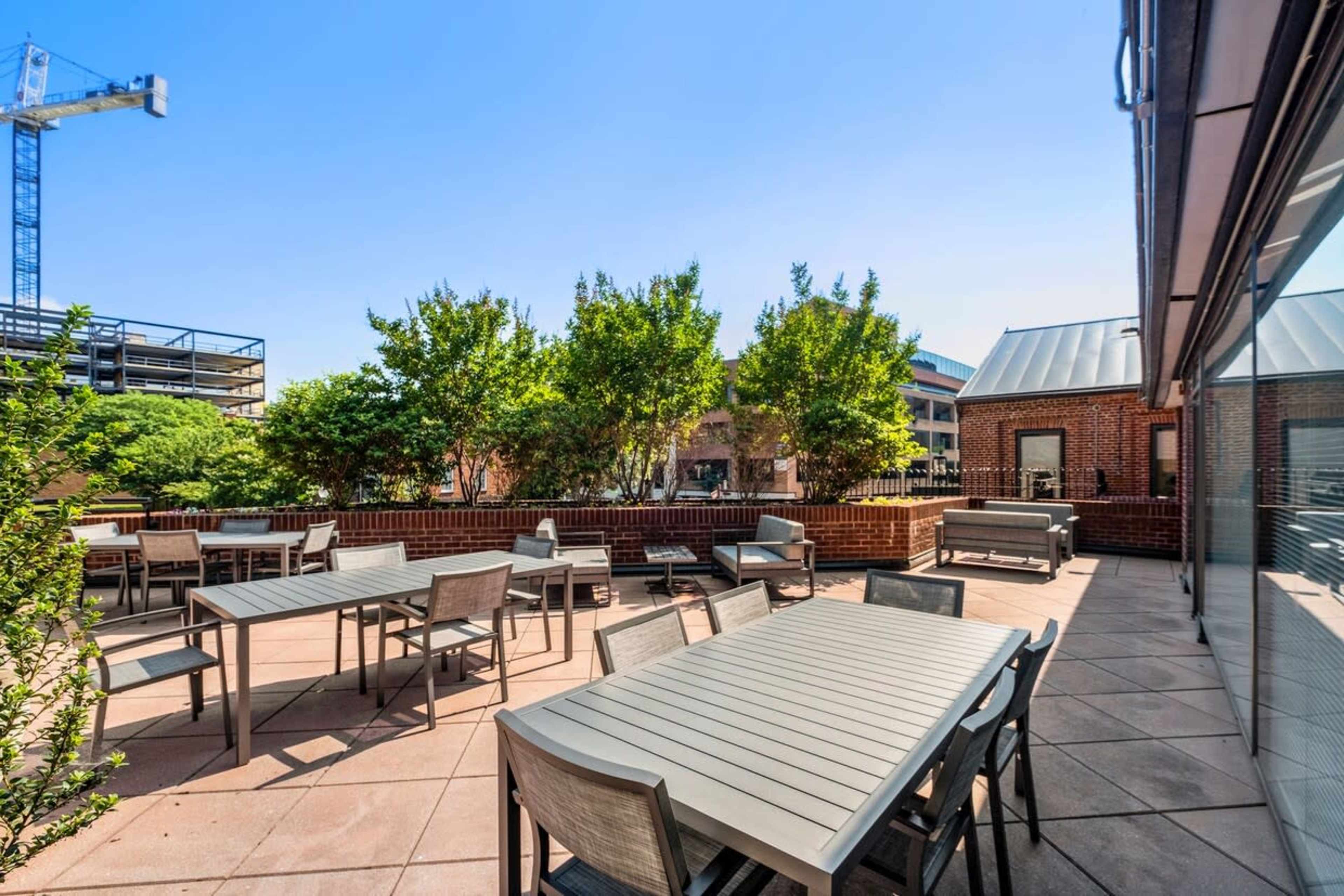 A patio area with several tables and chairs, surrounded by greenery and a clear blue sky.