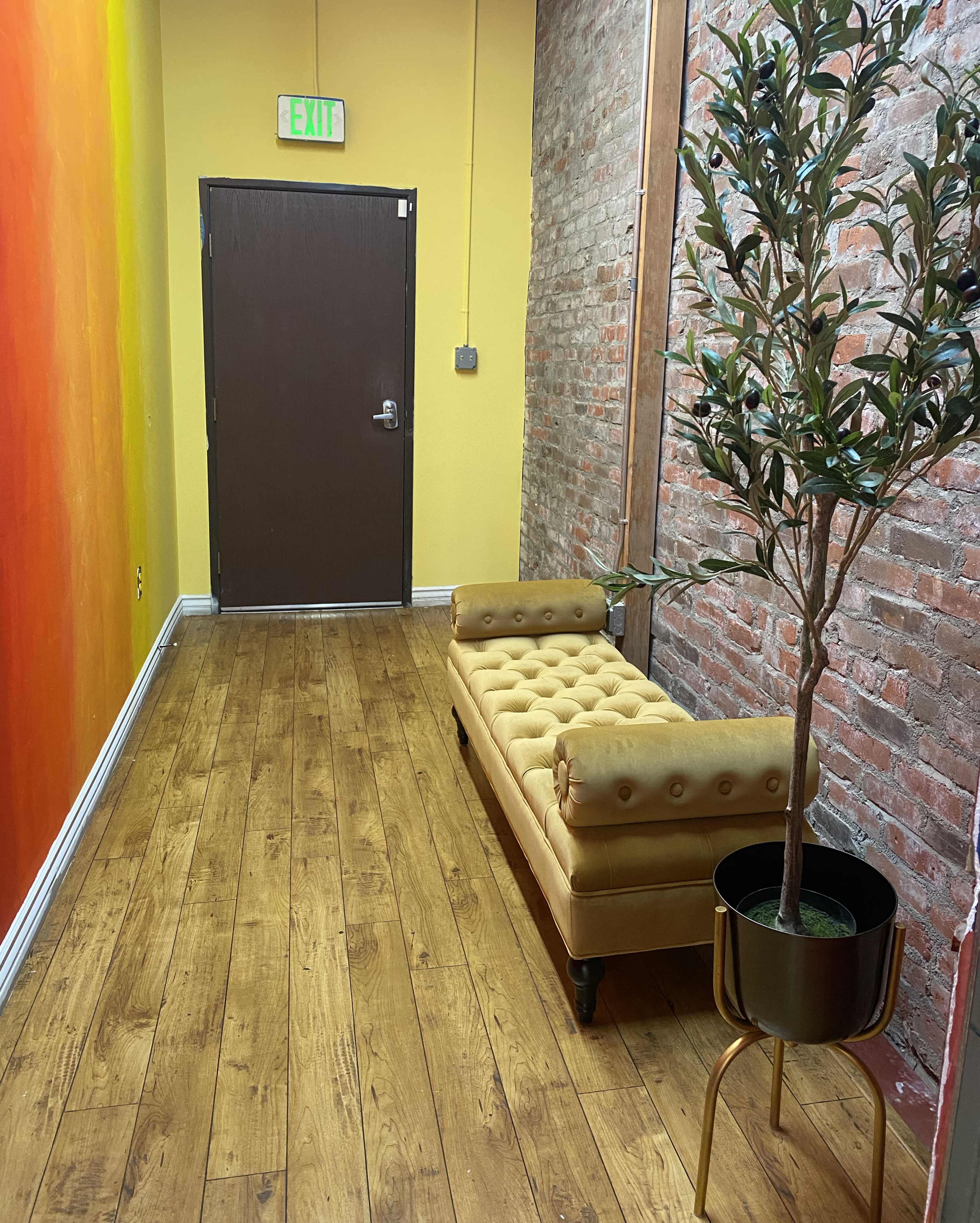 A narrow hallway features a yellow wall, a brick wall, a door at the end, a tufted yellow couch, and a potted plant.
