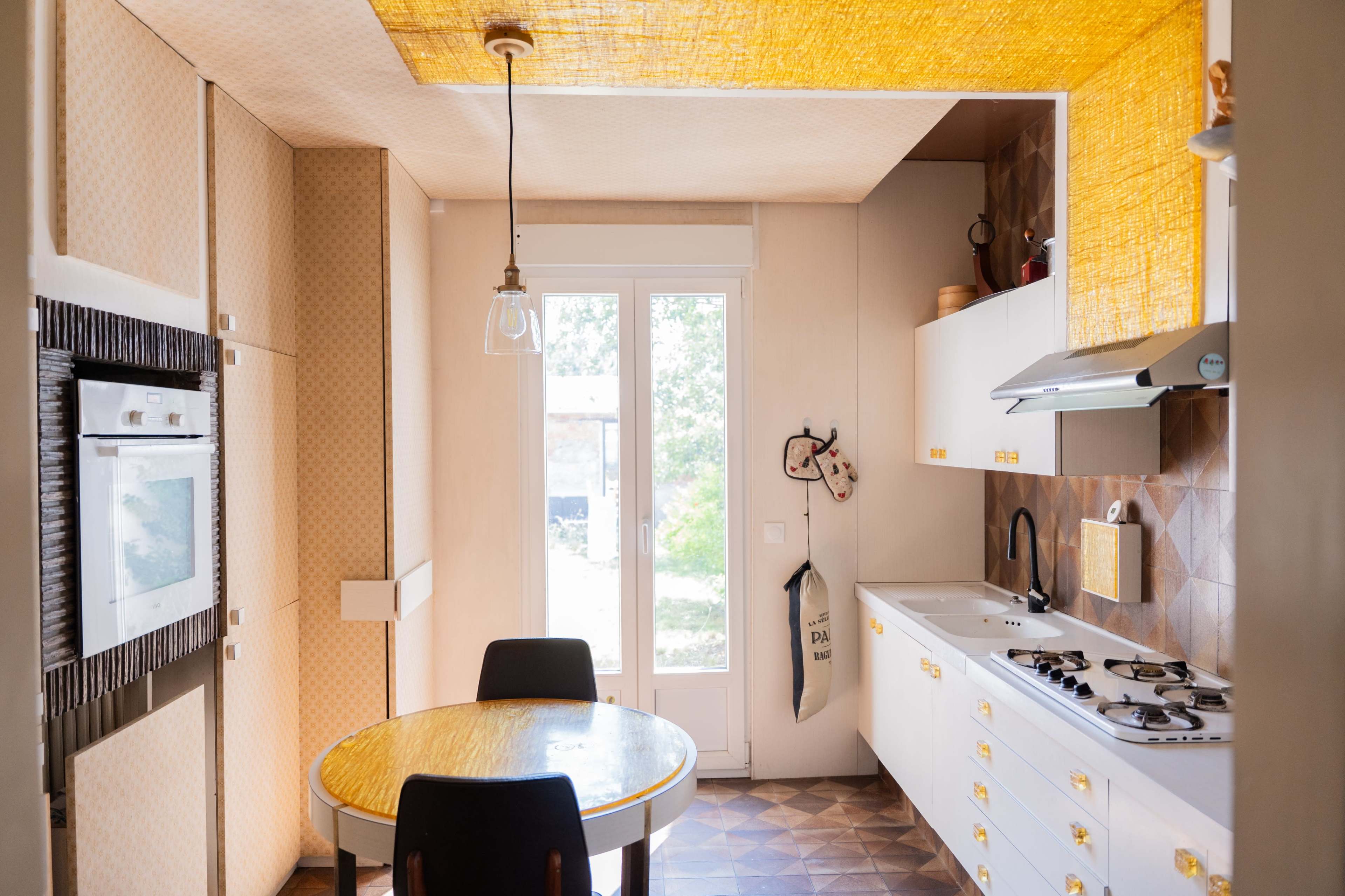 The image shows a bright kitchen with a round table, modern appliances, and a large window allowing natural light to enter.