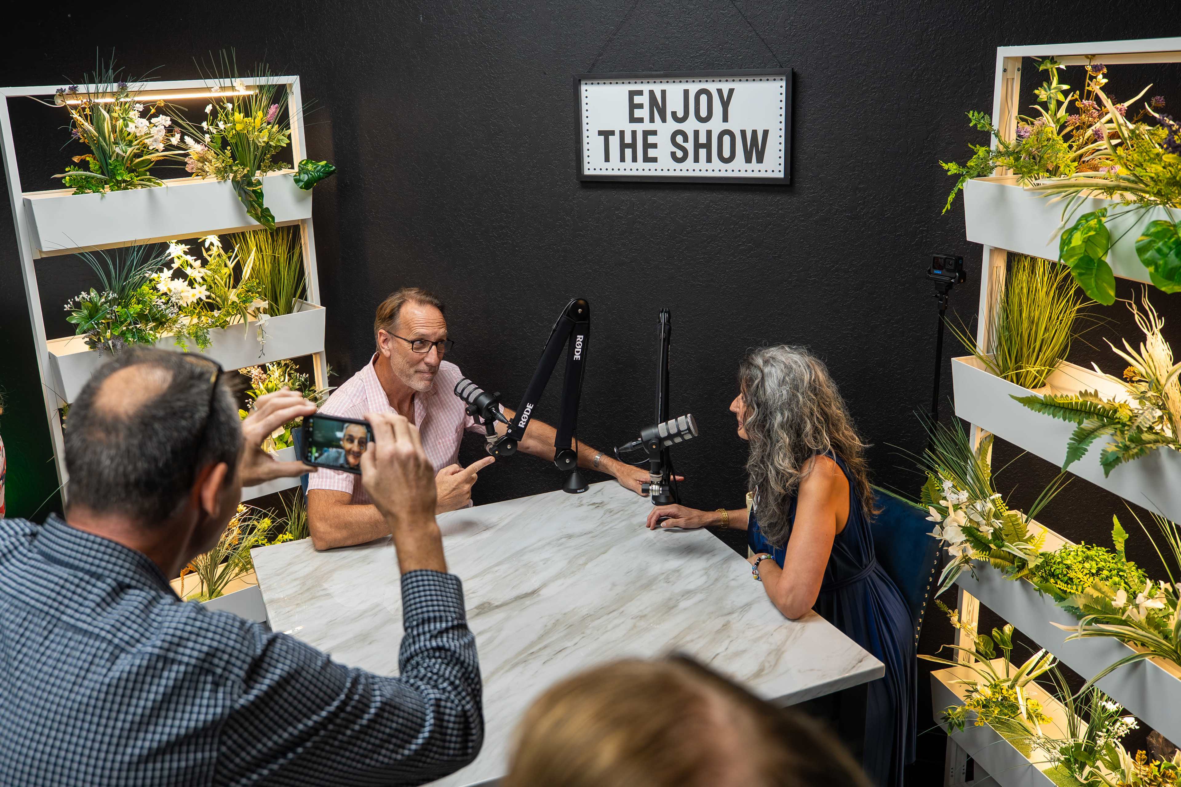 Two people are seated at a table with microphones in a decorated studio while a third person captures the moment with a camera.