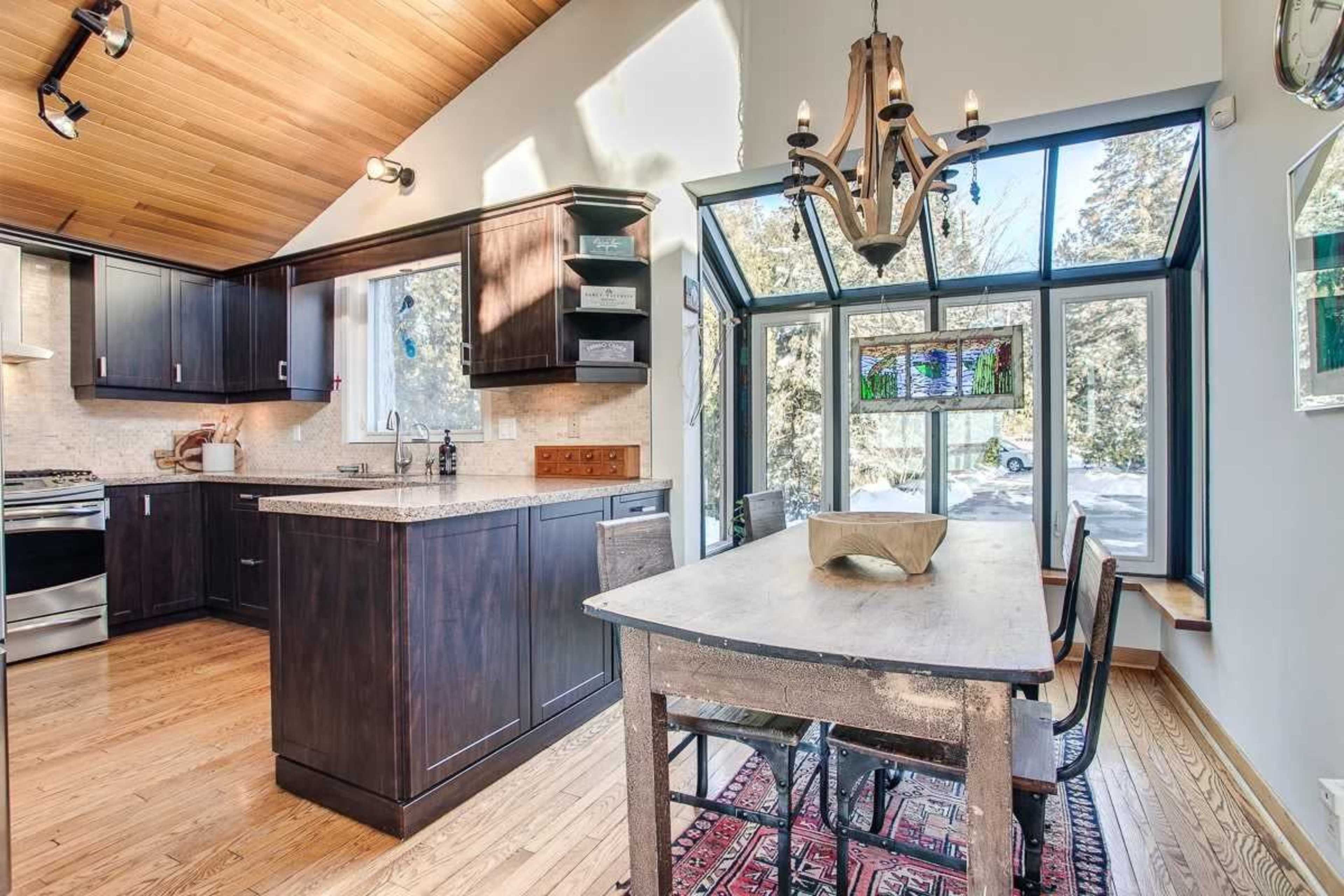 A modern kitchen featuring dark cabinetry, a granite countertop, and a dining area with a wooden table and chairs, illuminated by natural light from large windows.
