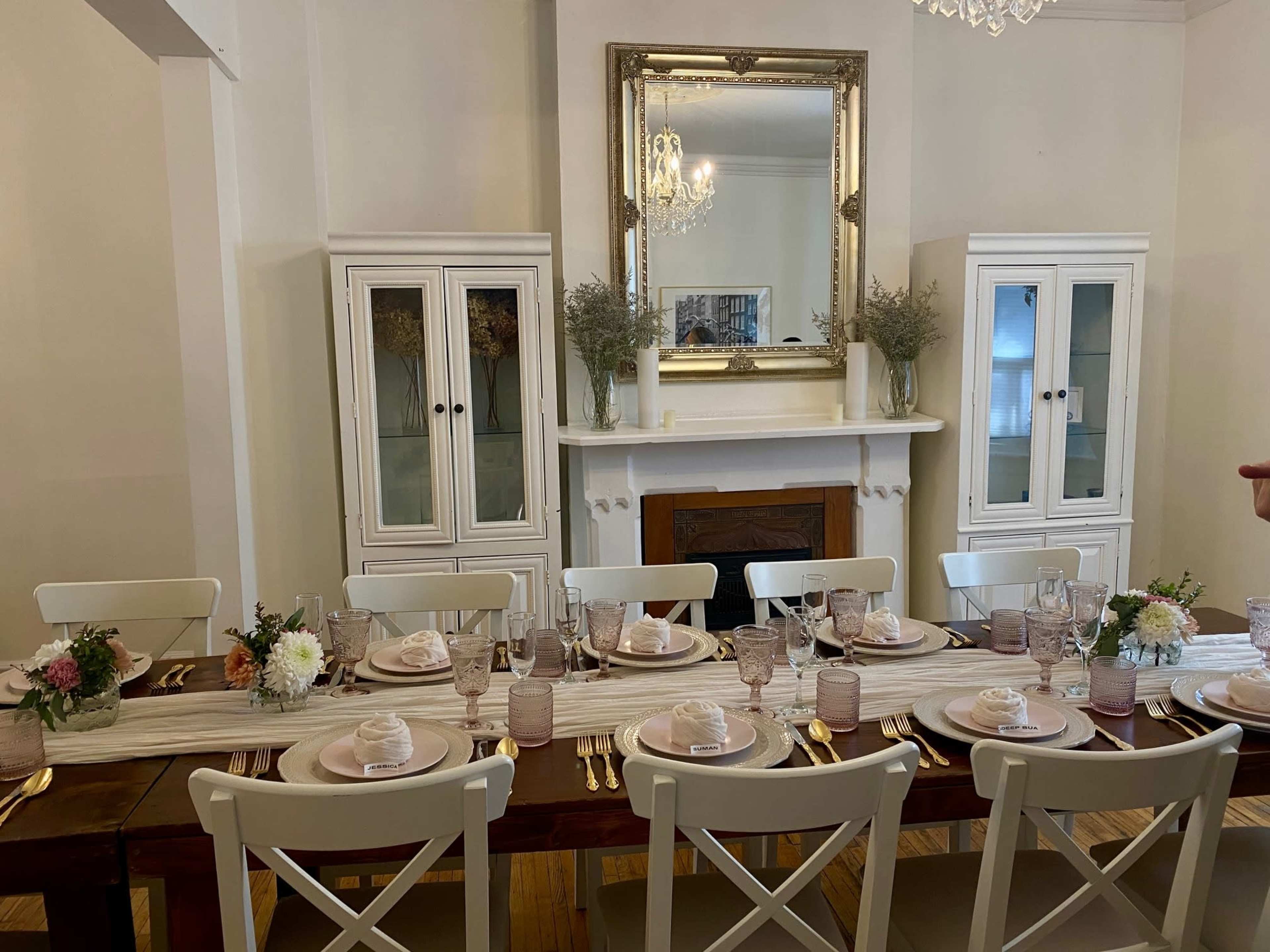 A long dining table is set with plates, glasses, and centerpieces, surrounded by white chairs in a room featuring a mirror and glass-front cabinets.