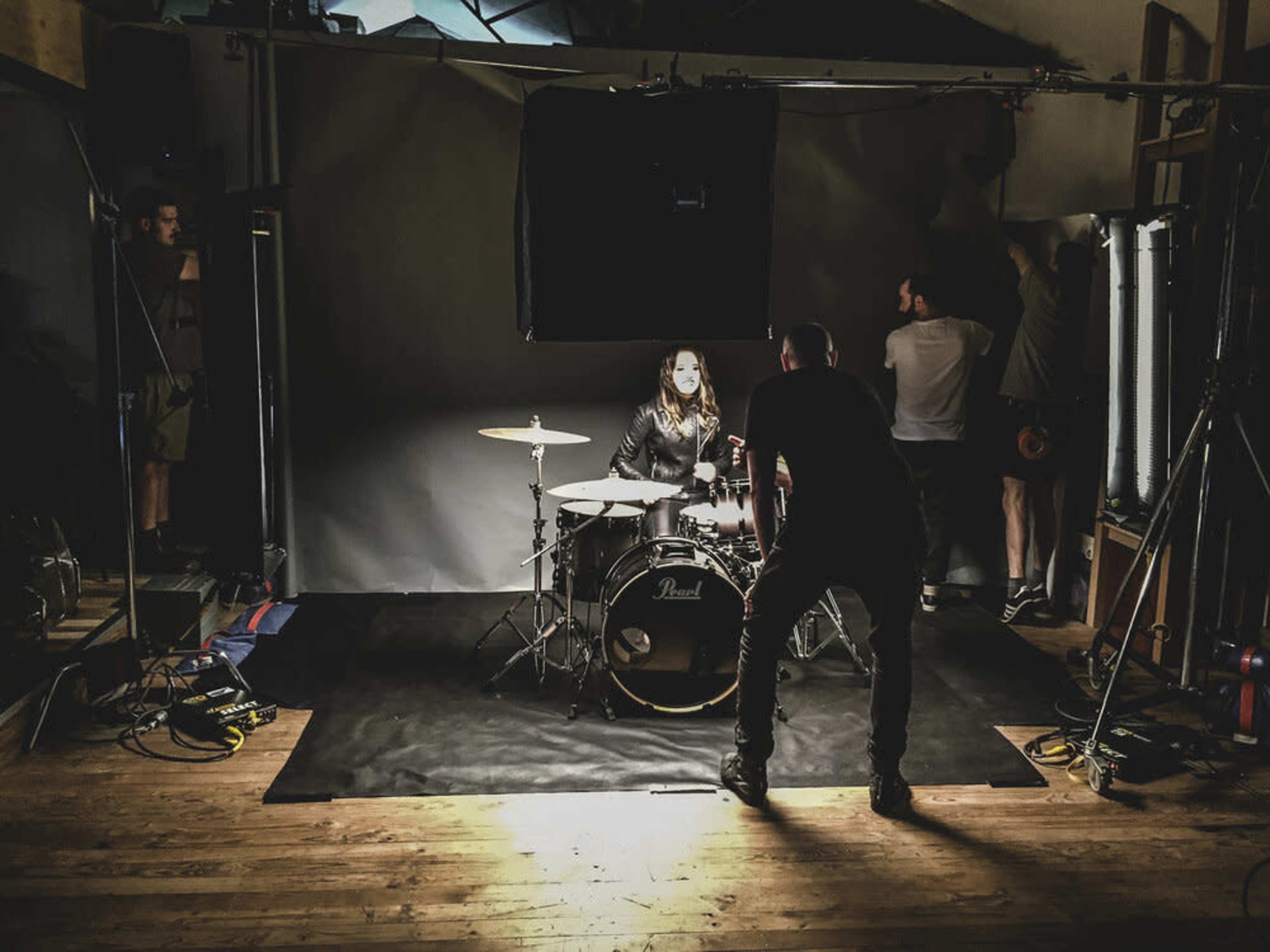 A drummer sits at a drum kit while a crew prepares the lighting and backdrop in a studio setting.