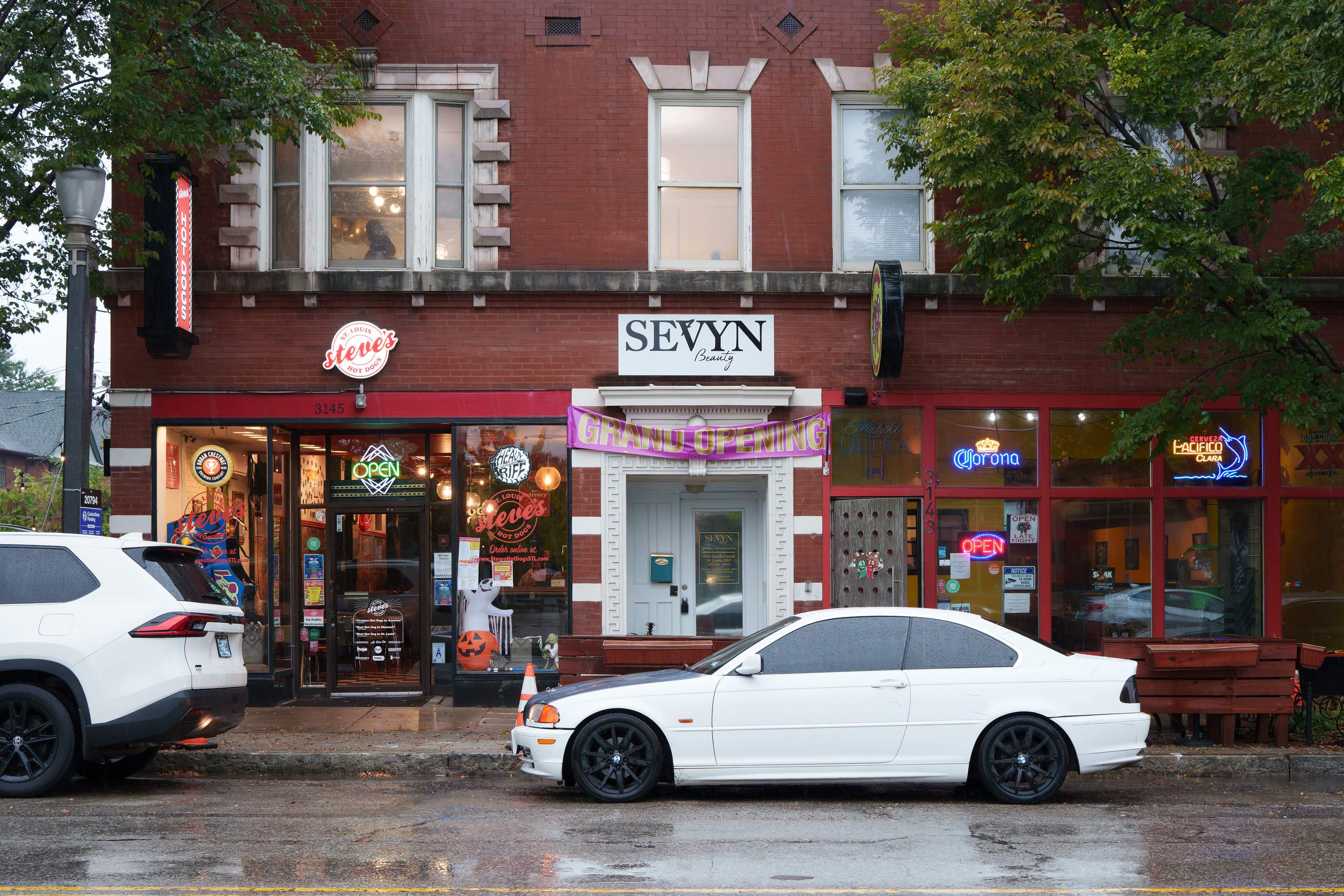 The image shows a storefront with several businesses, including a barbershop and a liquor store, located on a rainy street, with a white car parked in front.