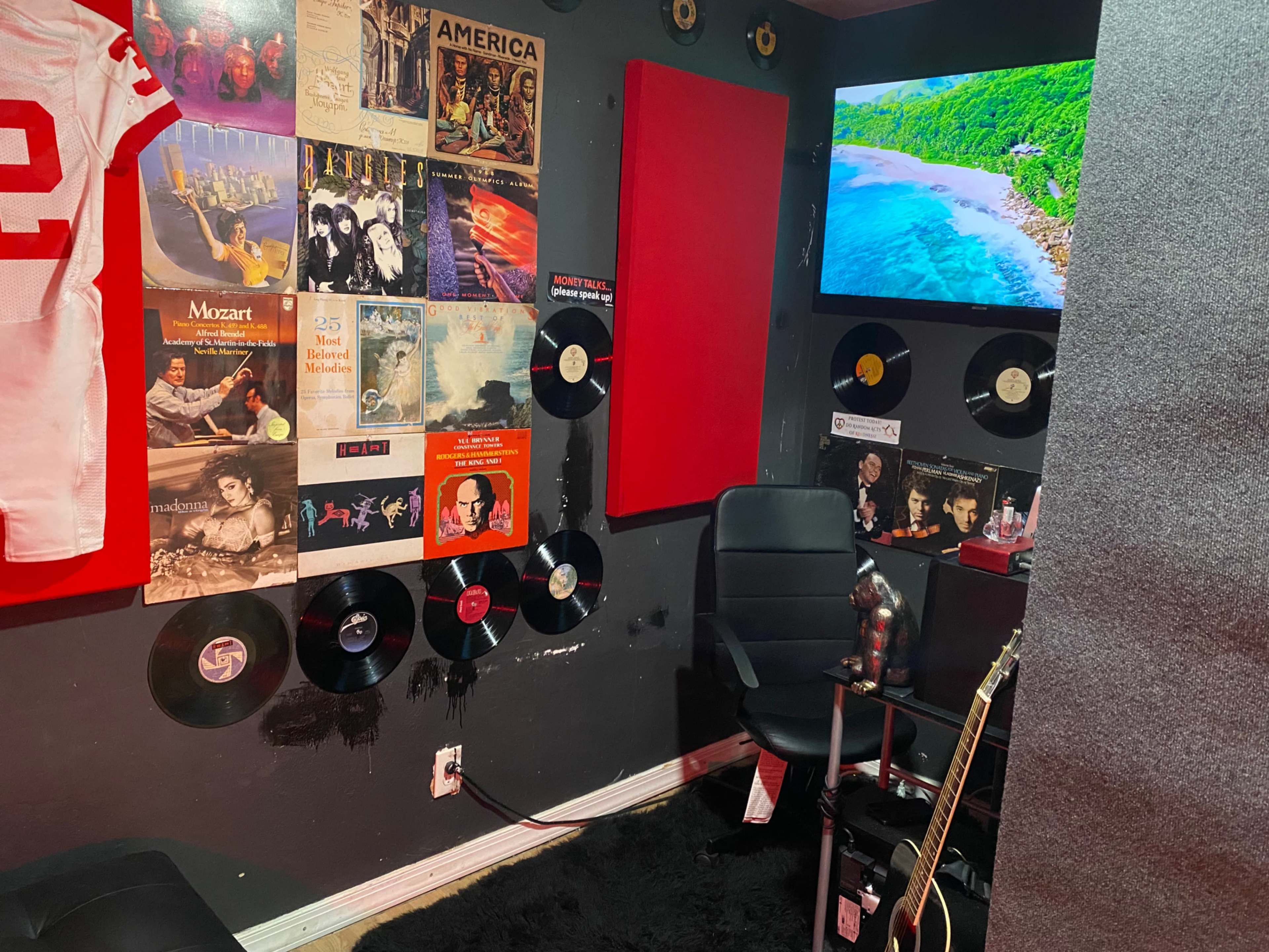 A small room featuring a collection of vinyl records on the walls, a red football jersey hanging, a chair, and a guitar positioned next to a television displaying a beach scene.