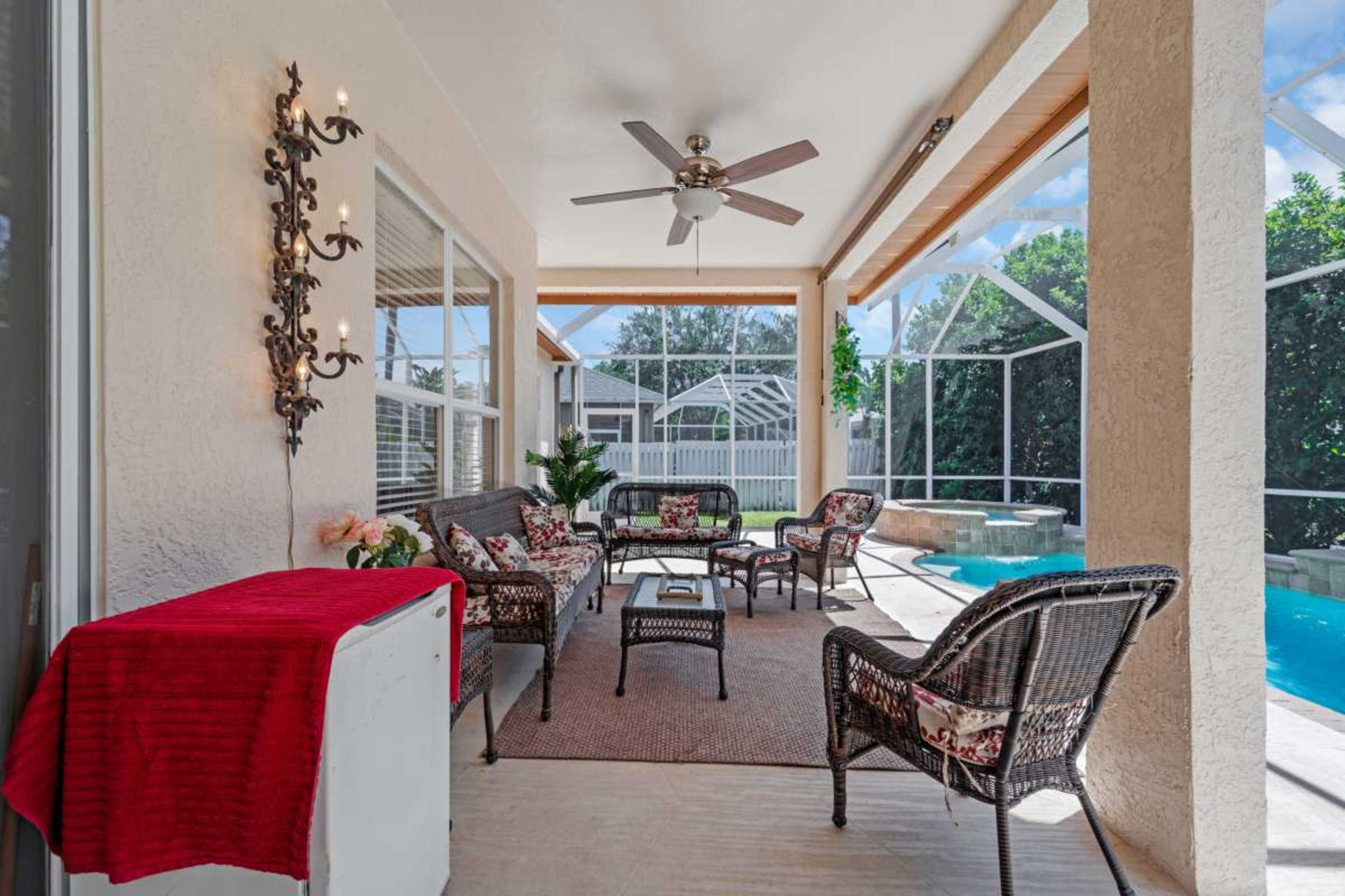 The image shows a covered patio with seating, a small table, and a view of a swimming pool through a screen enclosure.