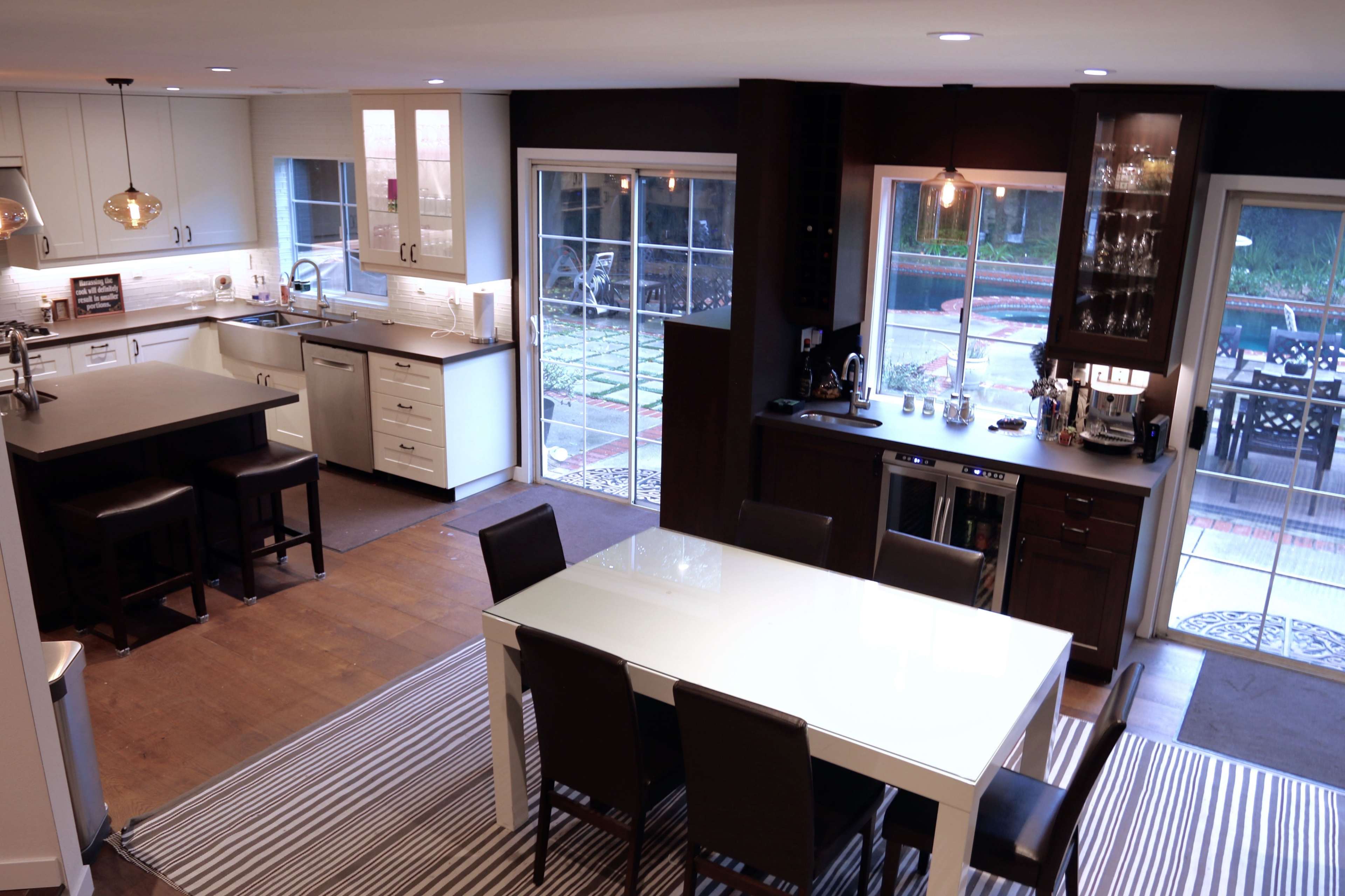 The image shows a modern kitchen and dining area with a large white table surrounded by black-chairs, complemented by a kitchen island and various appliances.