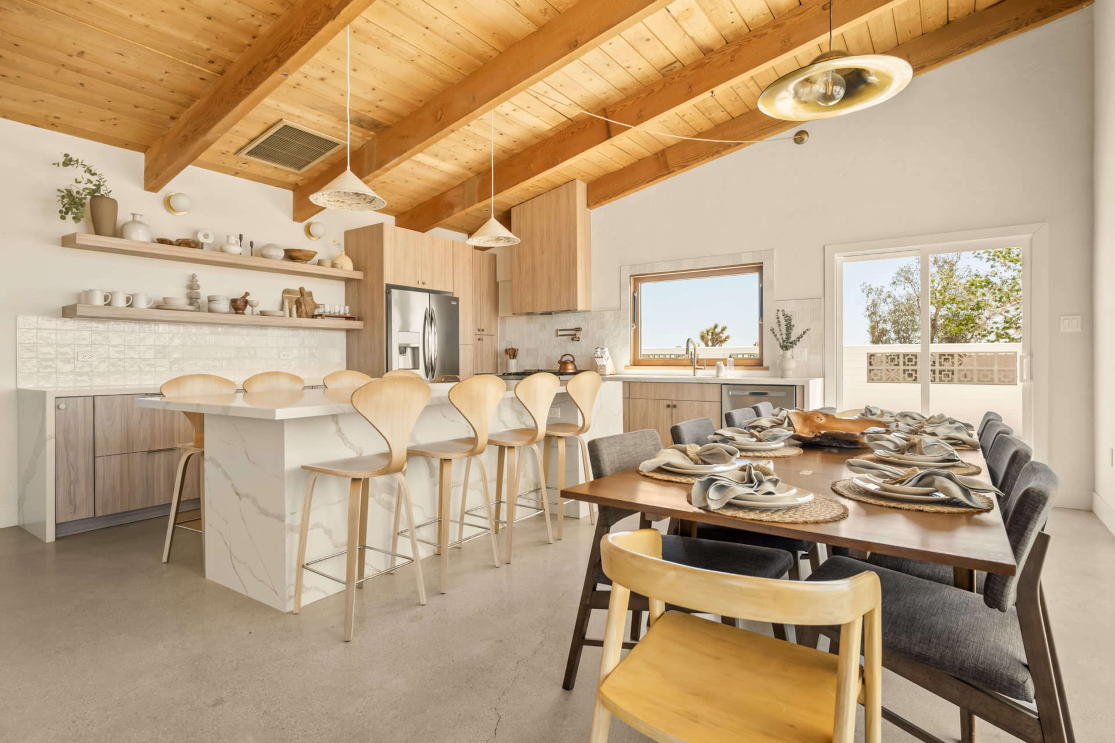 A modern kitchen features a large island with bar stools, a dining table set for a meal, and wooden beams on the ceiling.