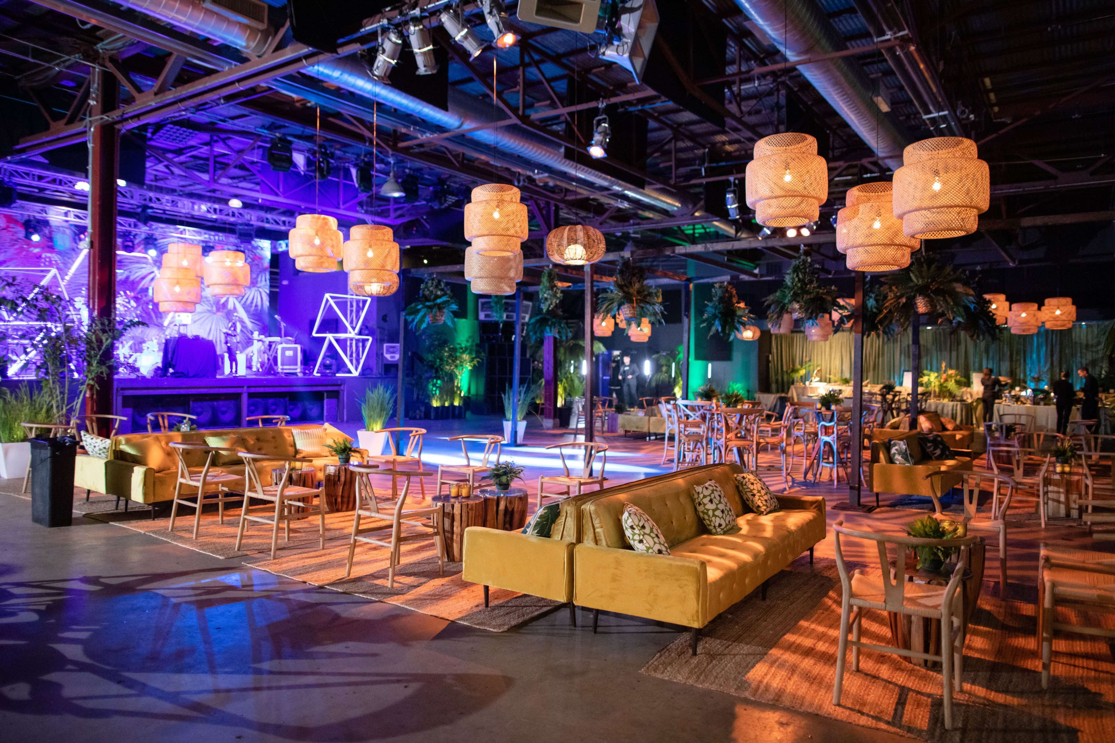 The image shows a spacious event venue with seated areas, decorative lighting, and tropical plants, creating a vibrant atmosphere.
