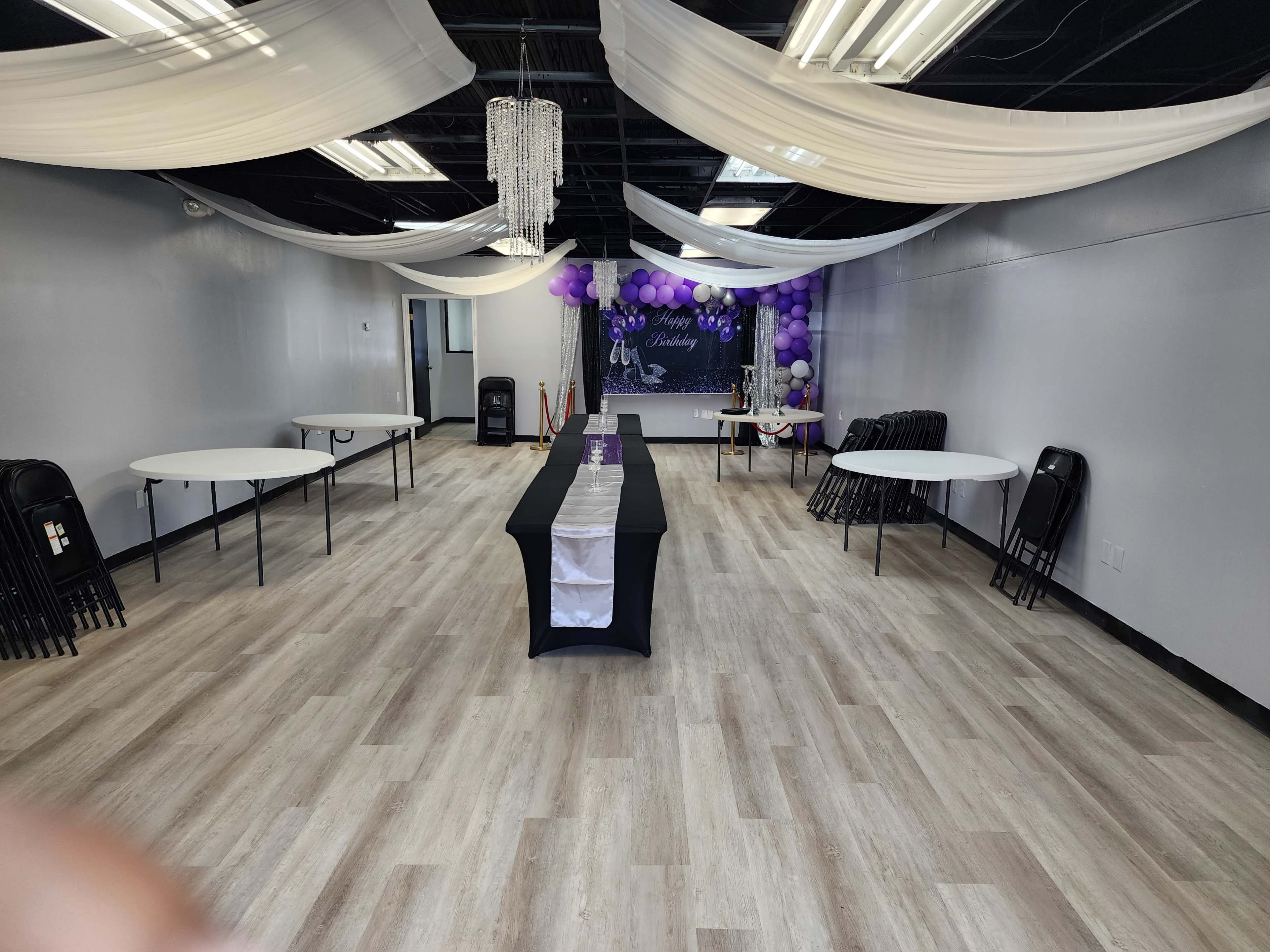 An empty event space with tables arranged around the room, adorned with purple balloons and draped fabric overhead.