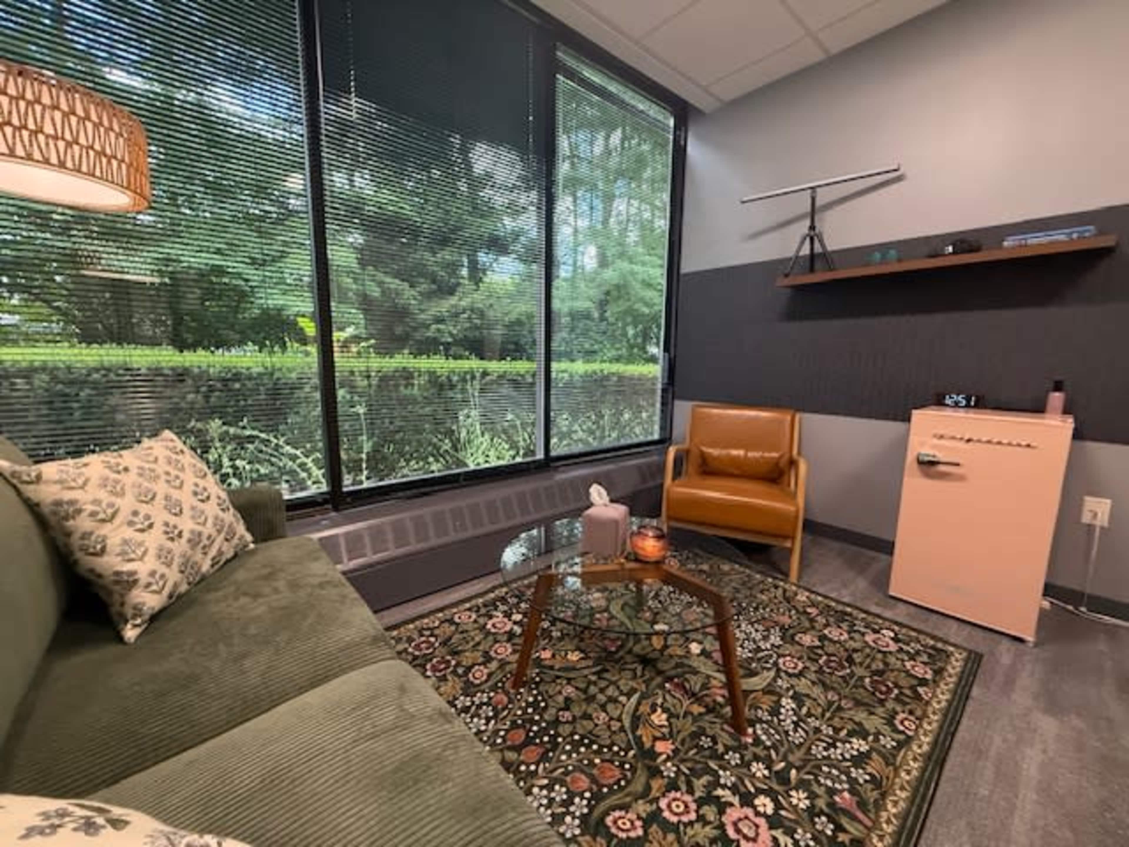 The image shows a small, cozy seating area with a green sofa, a brown armchair, a coffee table, and large windows that overlook greenery outside.