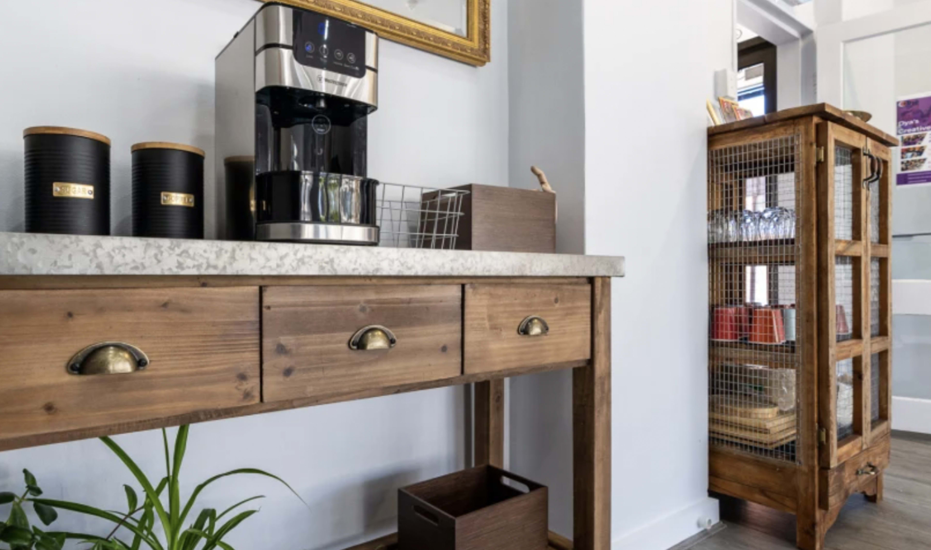 A wooden console table with drawers and a coffee maker is positioned against a wall, next to a wire-frame cabinet containing various items.