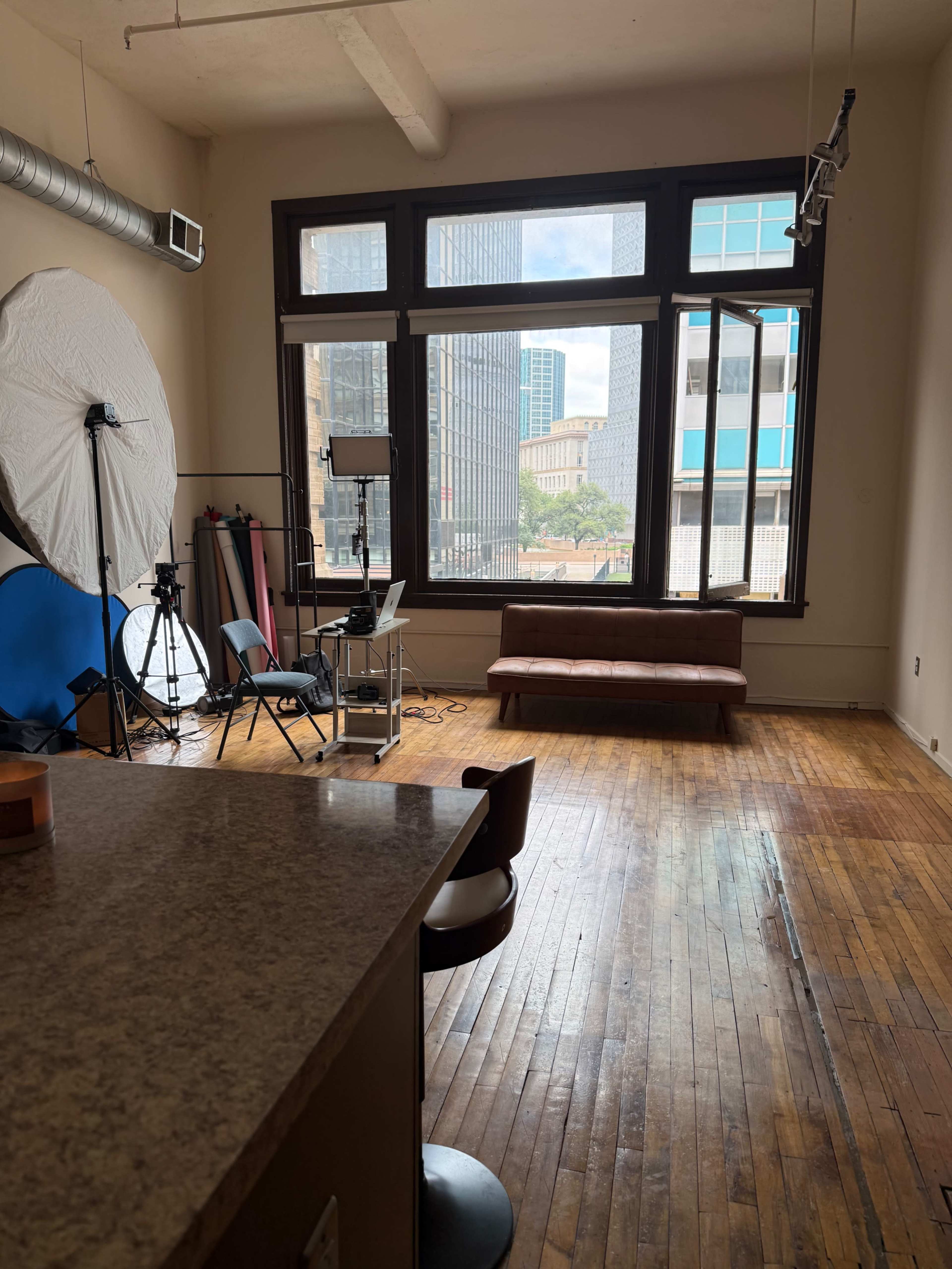 The image shows a spacious room with large windows letting in natural light, featuring a seating area, a backdrop set for photography, and a small desk with equipment.