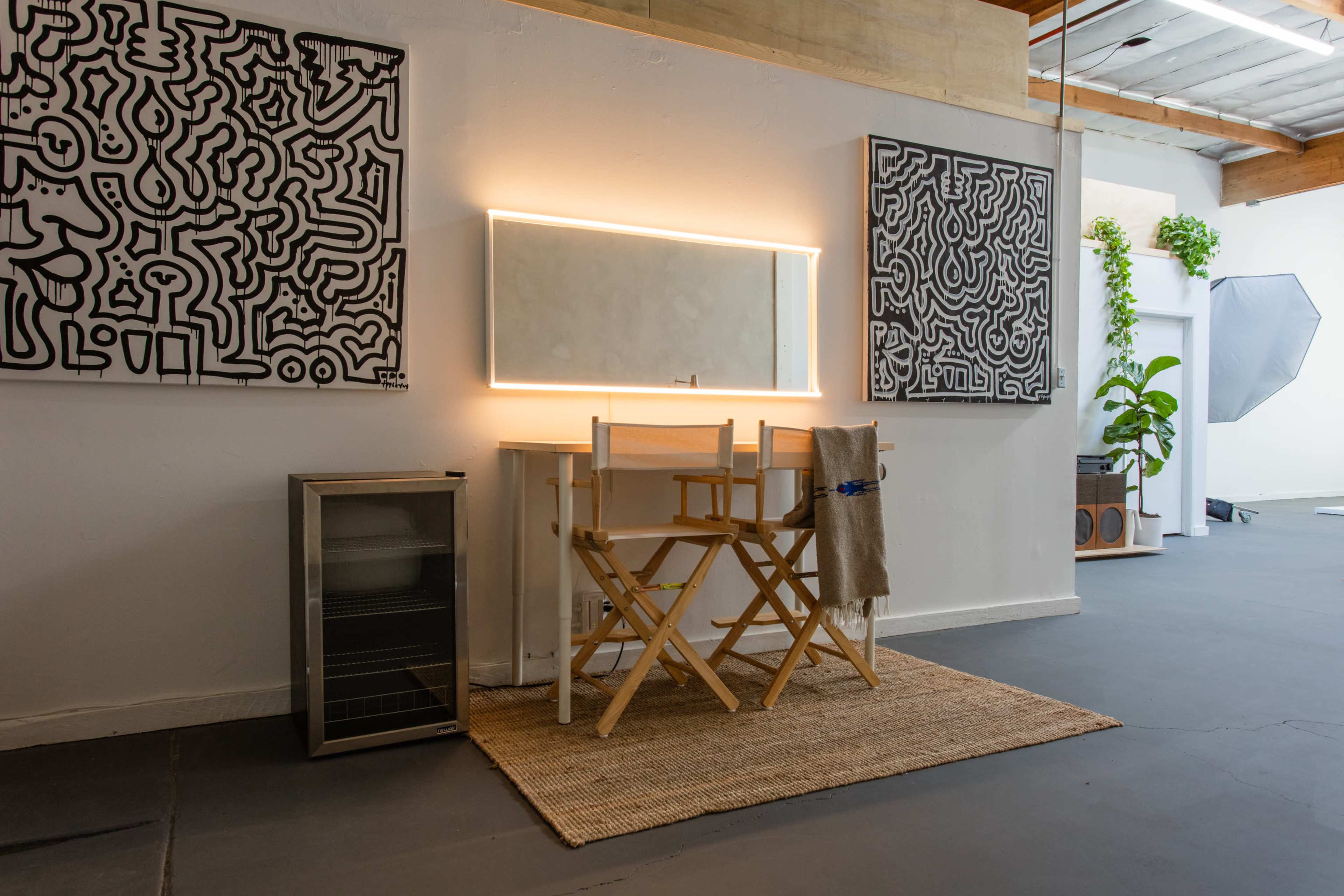 The image features a minimalist vanity area with a large mirror illuminated by LED lights, framed by two abstract black-and-white artworks, a small refrigerator, and a woven rug on the floor.