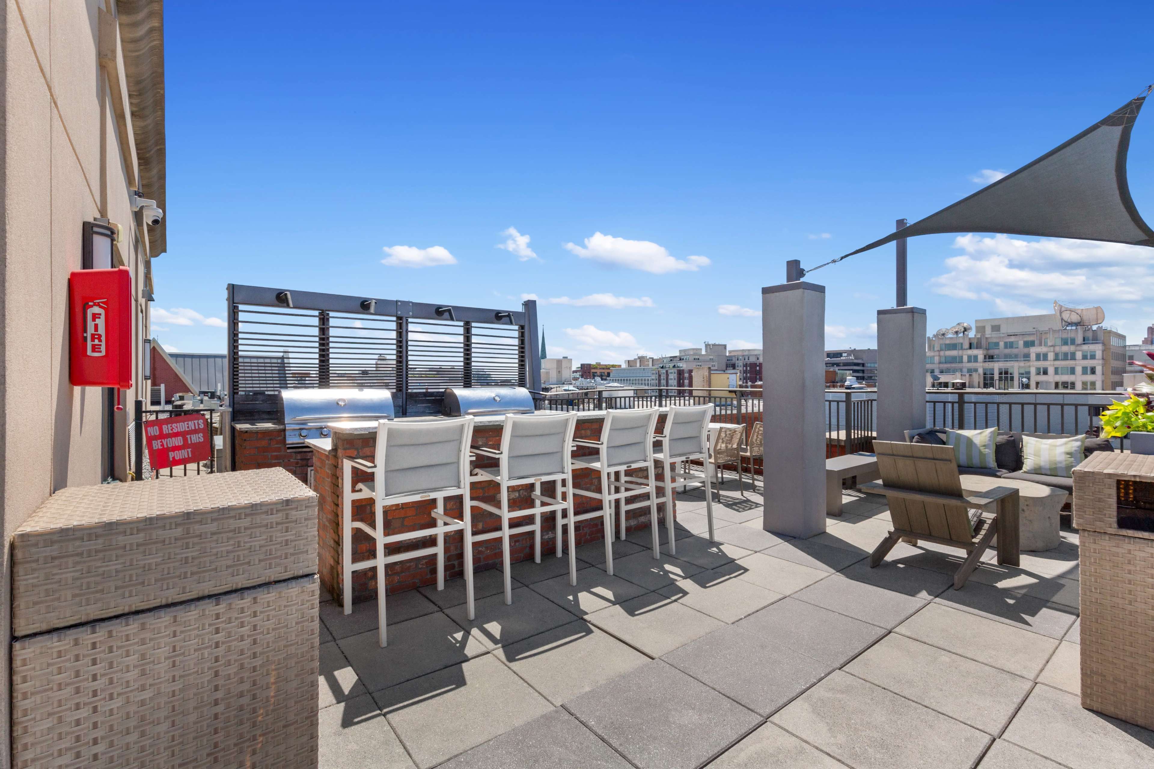 The image shows a rooftop space featuring a grill station, bar-style seating, and a shaded seating area with a clear blue sky in the background.