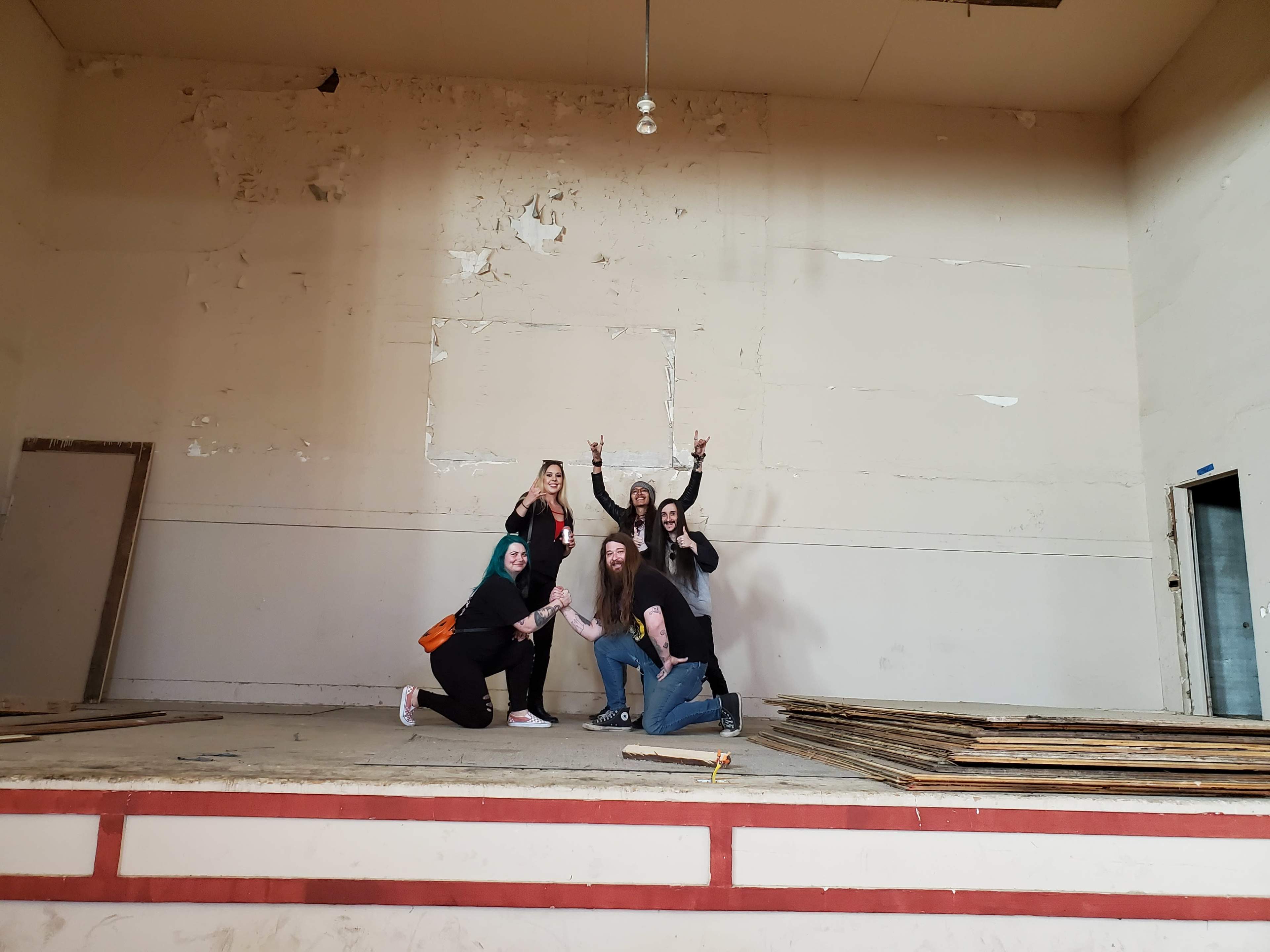 A group of five individuals poses on a wooden stage in an empty room with bare walls.