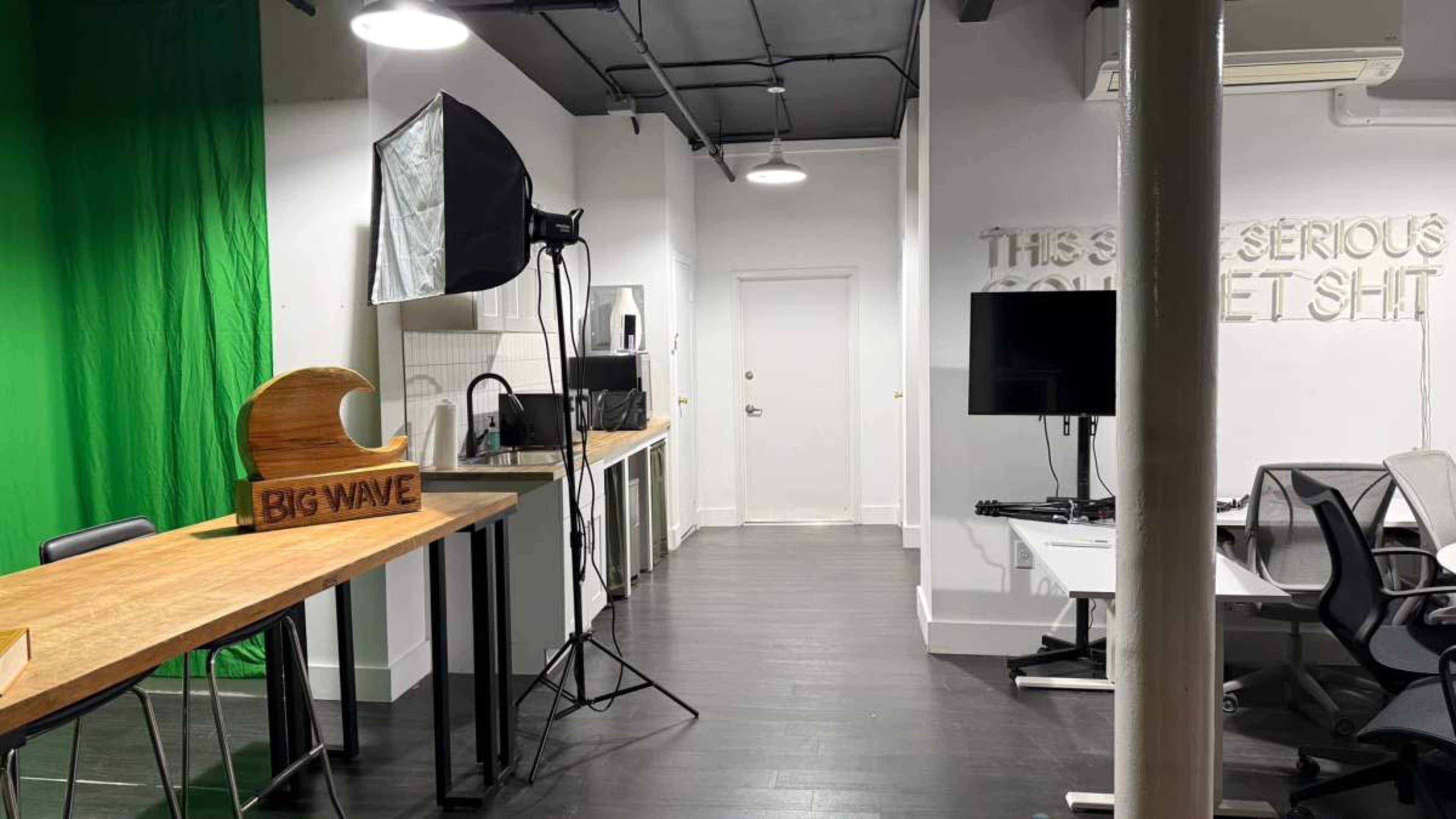 The image shows a modern studio space with a green screen, lighting equipment, a wooden table with a "BIG WAVE" sign, and multiple desks in a well-lit environment.