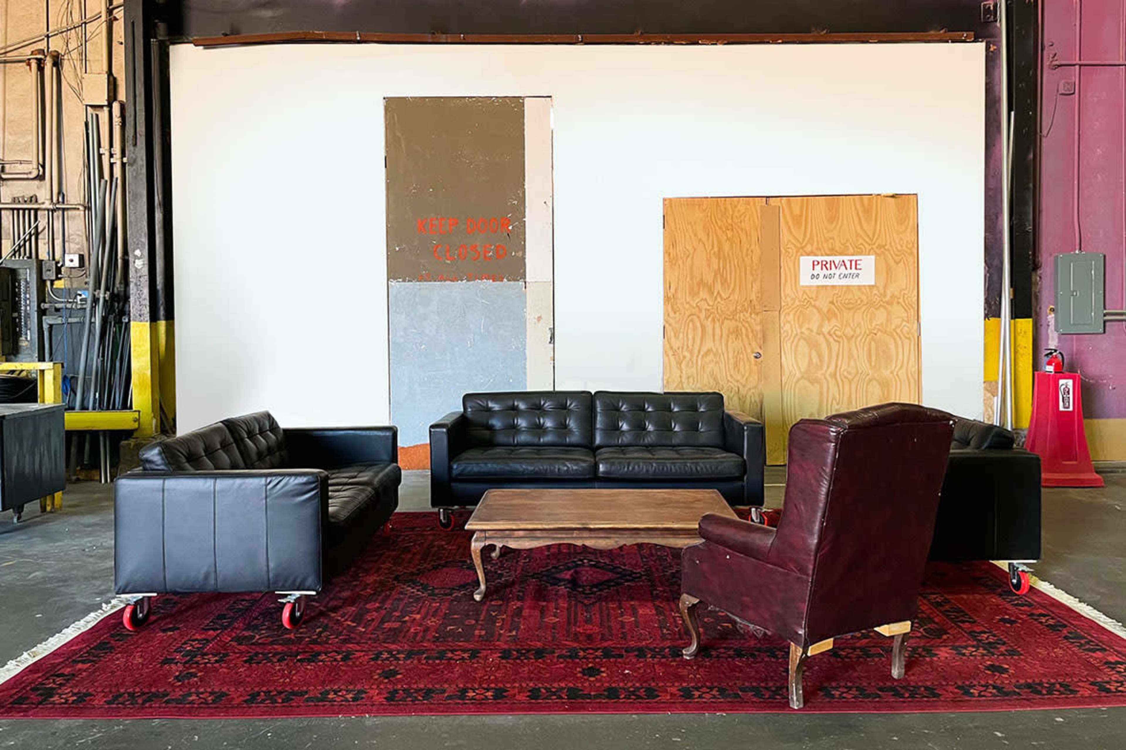 The image shows a seating arrangement comprising three black leather sofas, a wooden coffee table, and a burgundy armchair, all situated on a red patterned rug in an industrial setting.