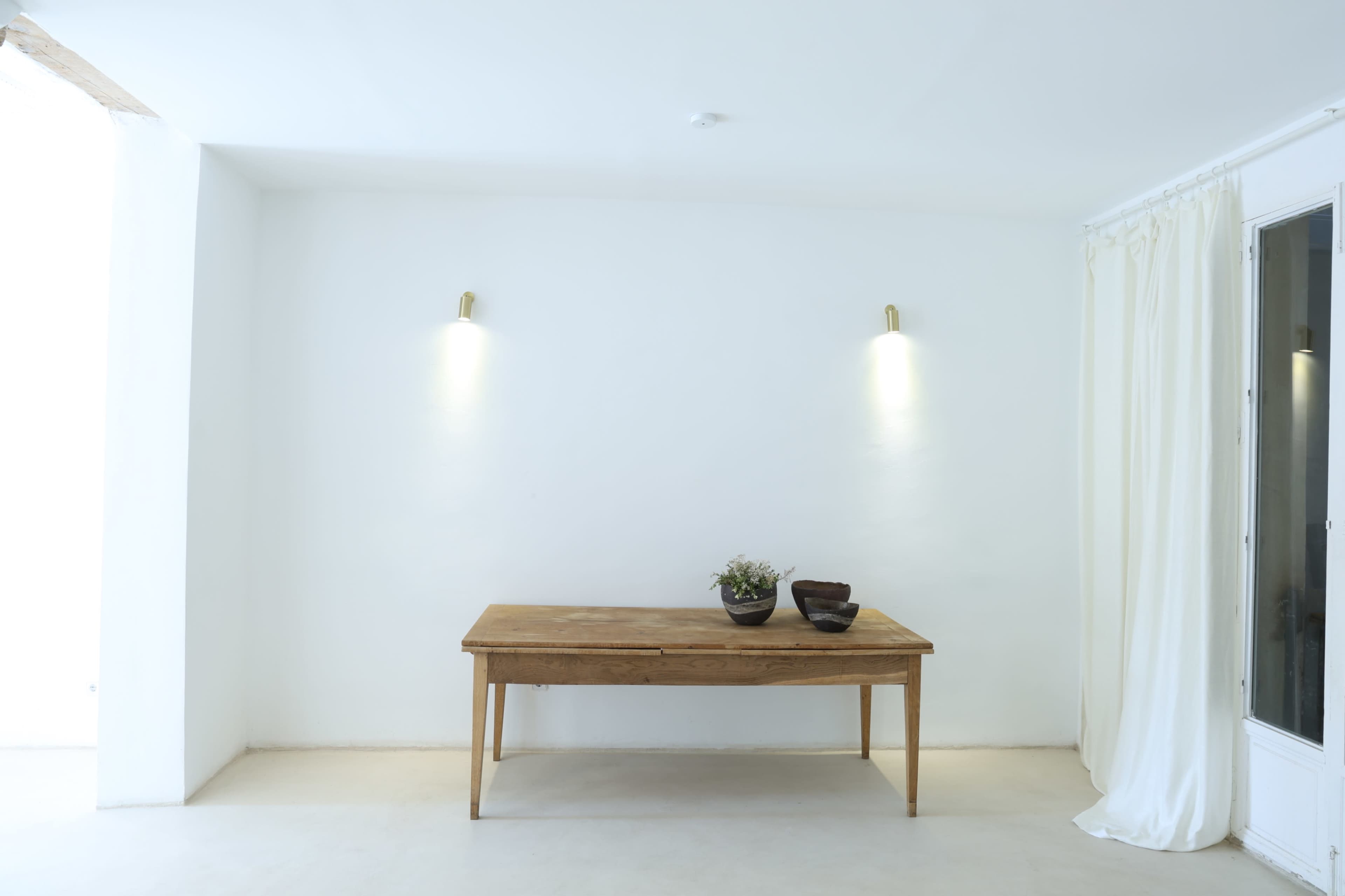 The image shows a minimalist interior with a wooden table, two wall sconces, and a white curtain in a well-lit space.
