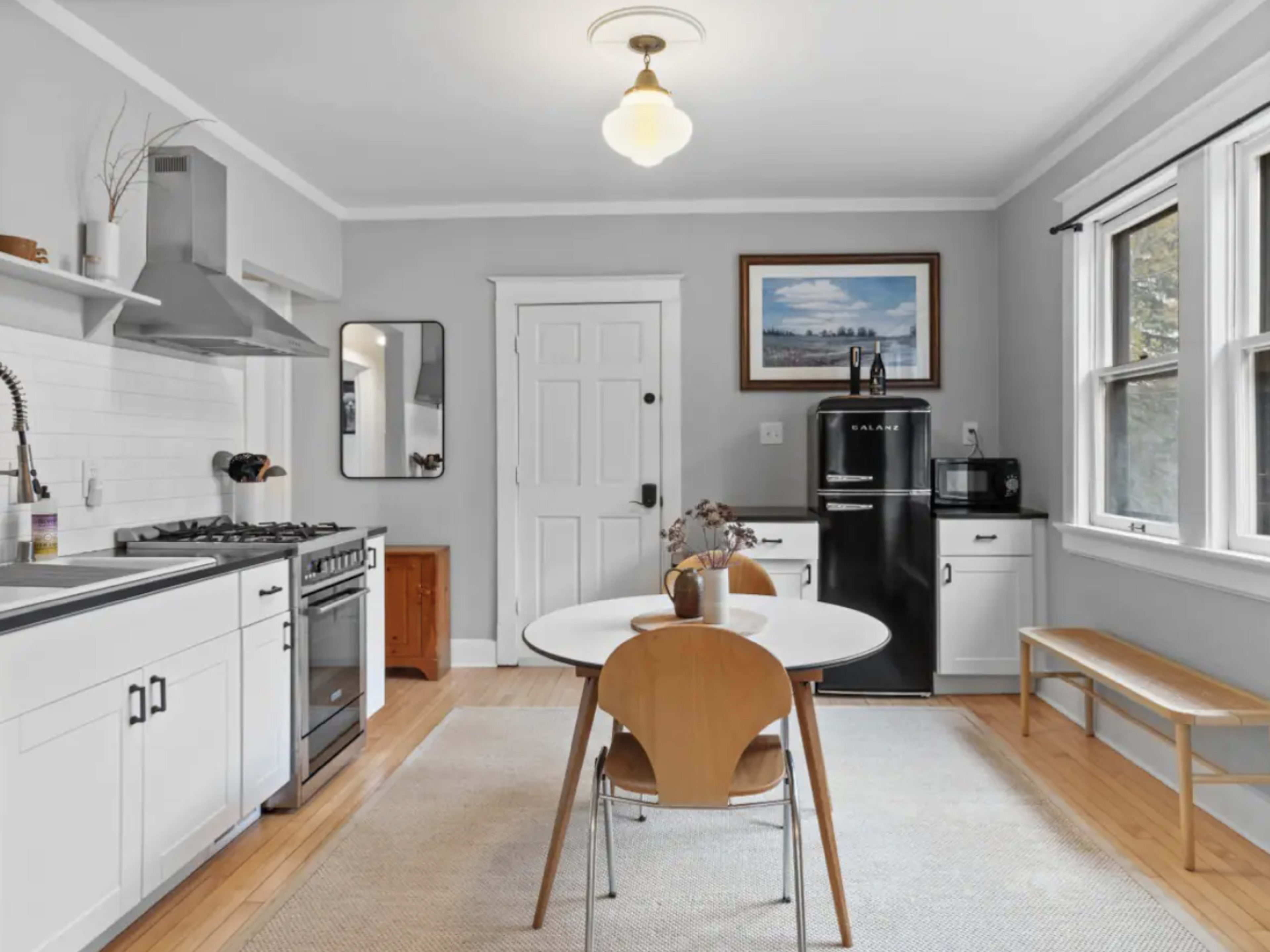 A modern kitchen features a circular table with wooden chairs, a black refrigerator, and stainless steel appliances against light gray walls.