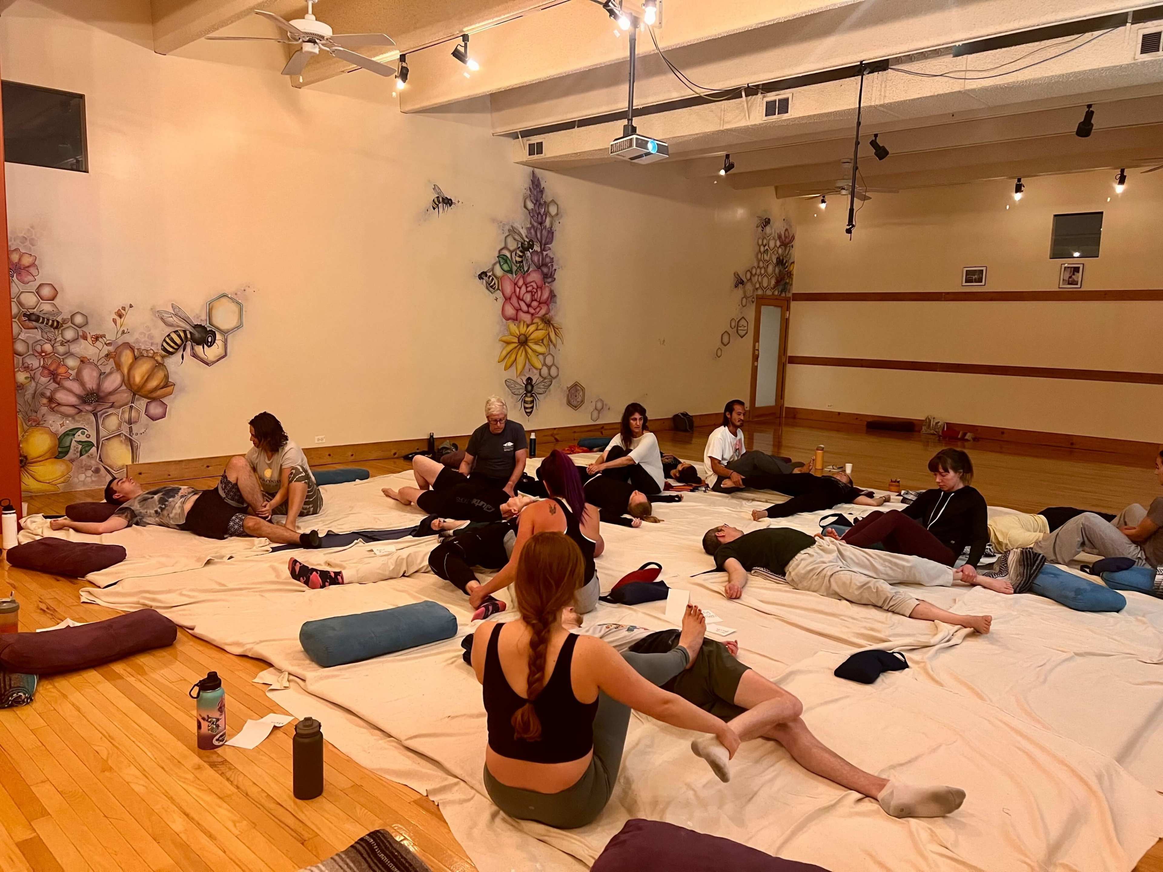 A group of people are lying on mats and cushions in a spacious room decorated with colorful murals.