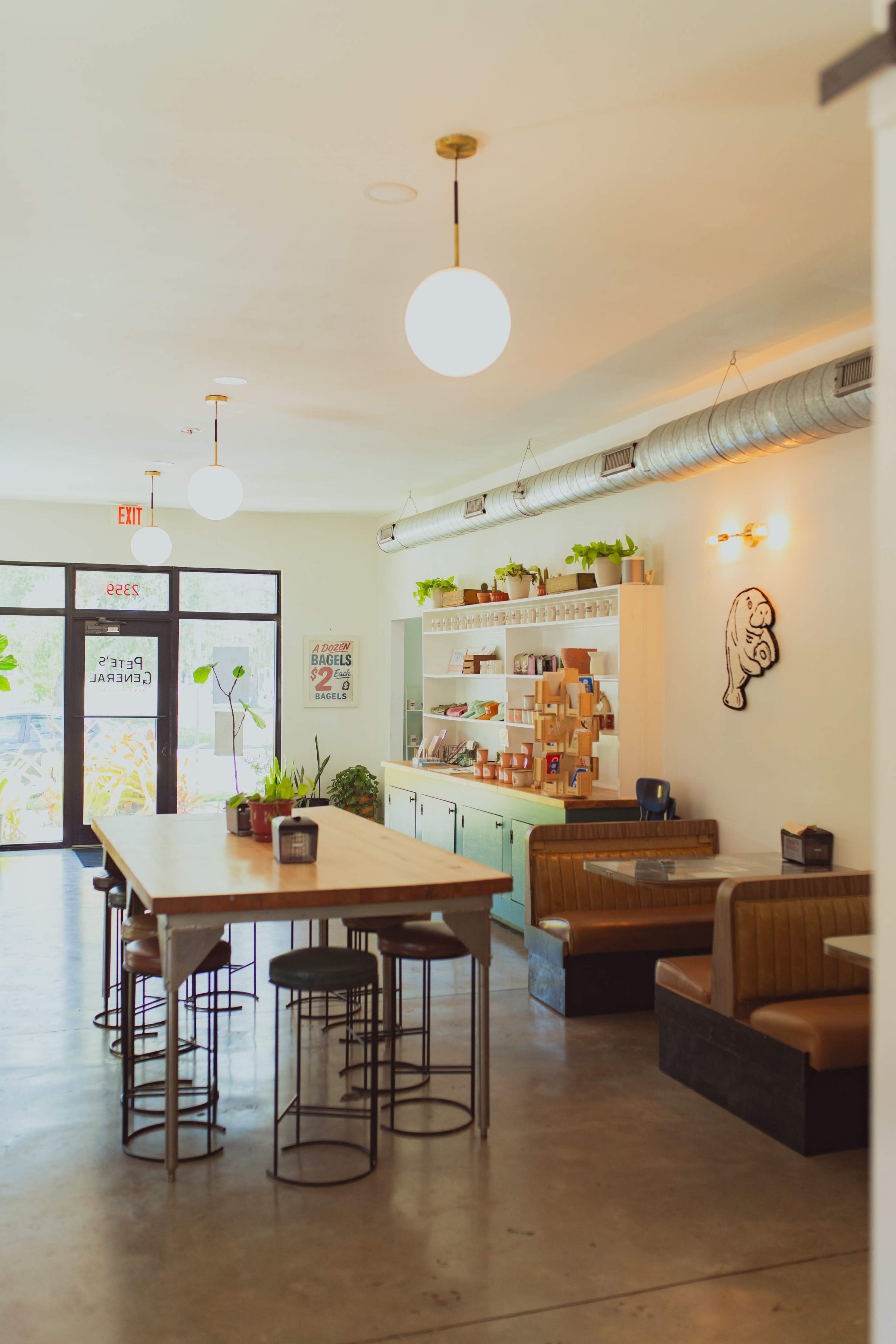 The image shows a modern cafe interior featuring a long wooden table with metal stools, cozy seating along the walls, and large windows allowing natural light to enter.