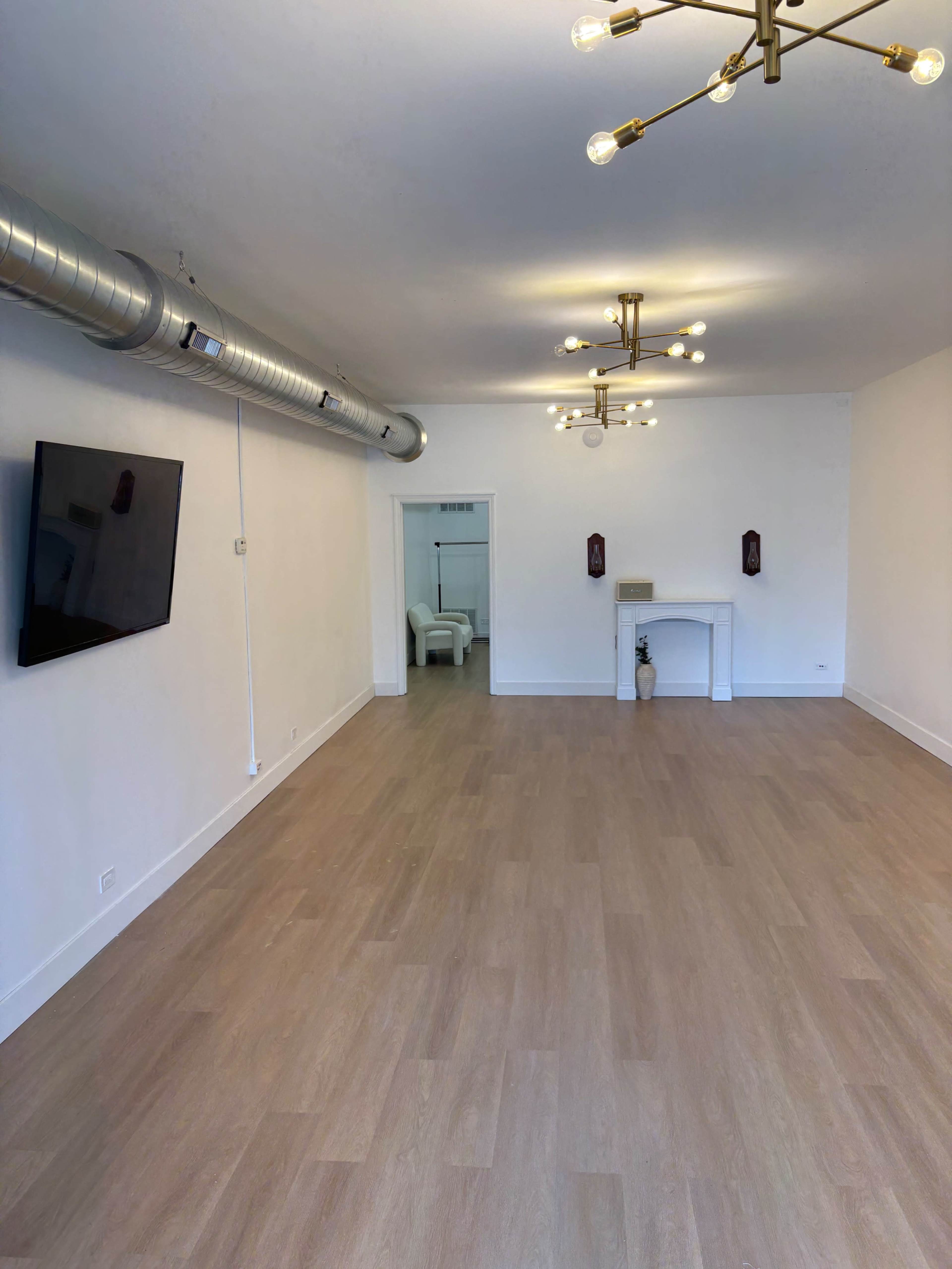 The image shows a spacious, minimally furnished room with light wood flooring, a wall-mounted television, and modern light fixtures.