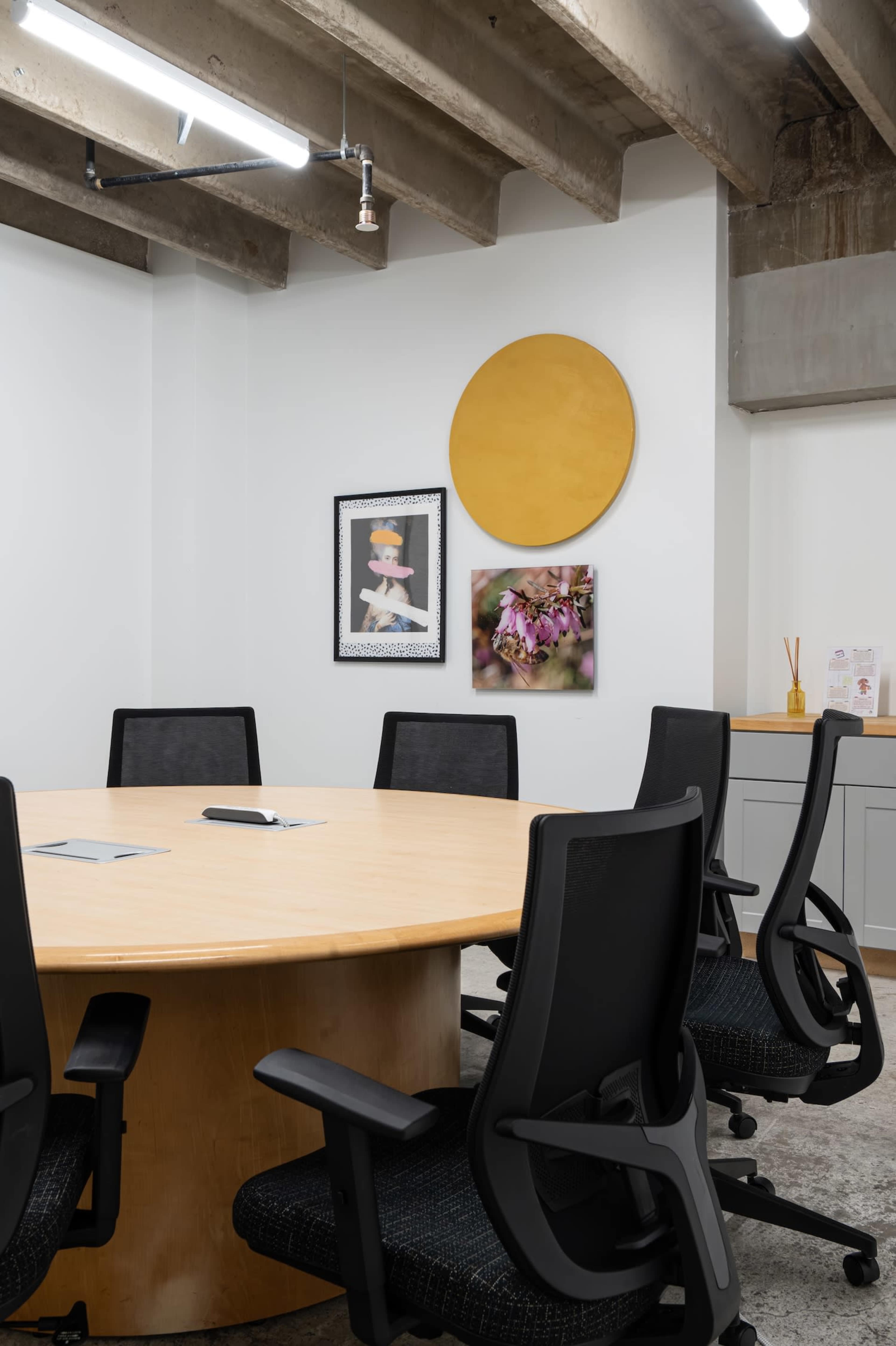 The image shows a modern conference room with a round wooden table surrounded by black office chairs, featuring framed artwork on the walls and a large circular artwork above the table.