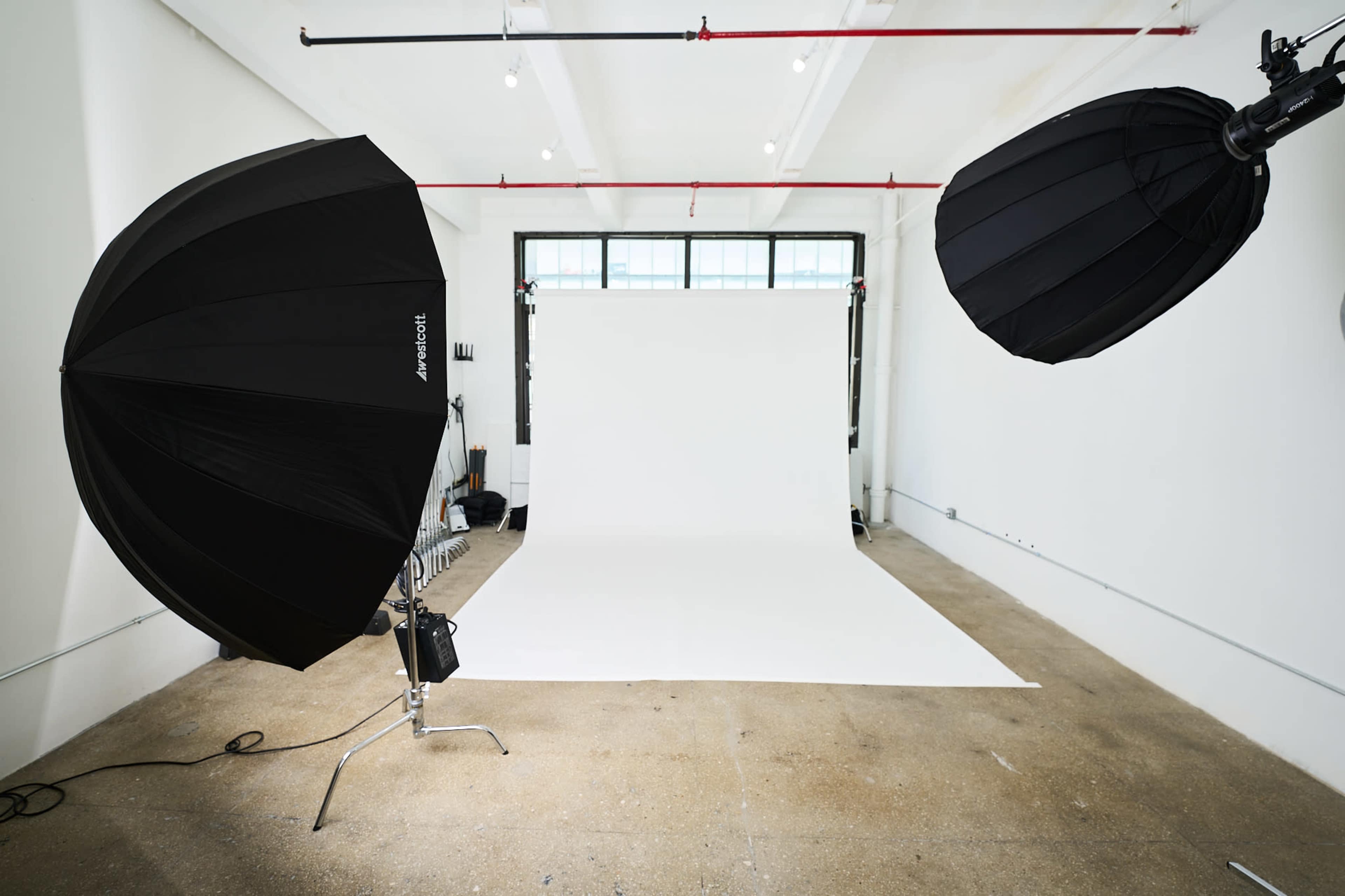 The image shows a photography studio with two large softbox lights positioned on either side of a plain white backdrop and a concrete floor.