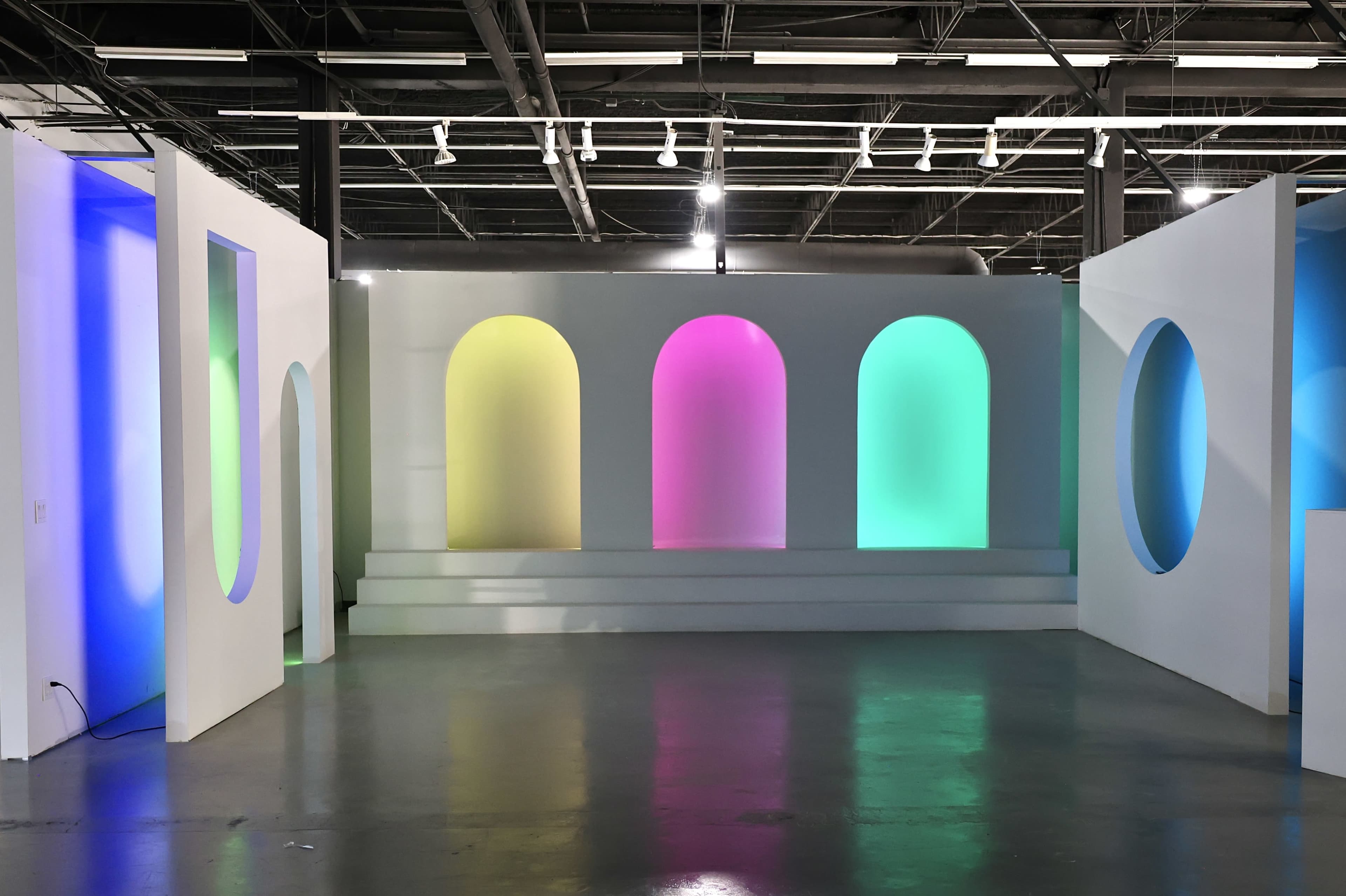 The image shows a large, modern indoor space with white walls and colorful lighting in arch-shaped alcoves.