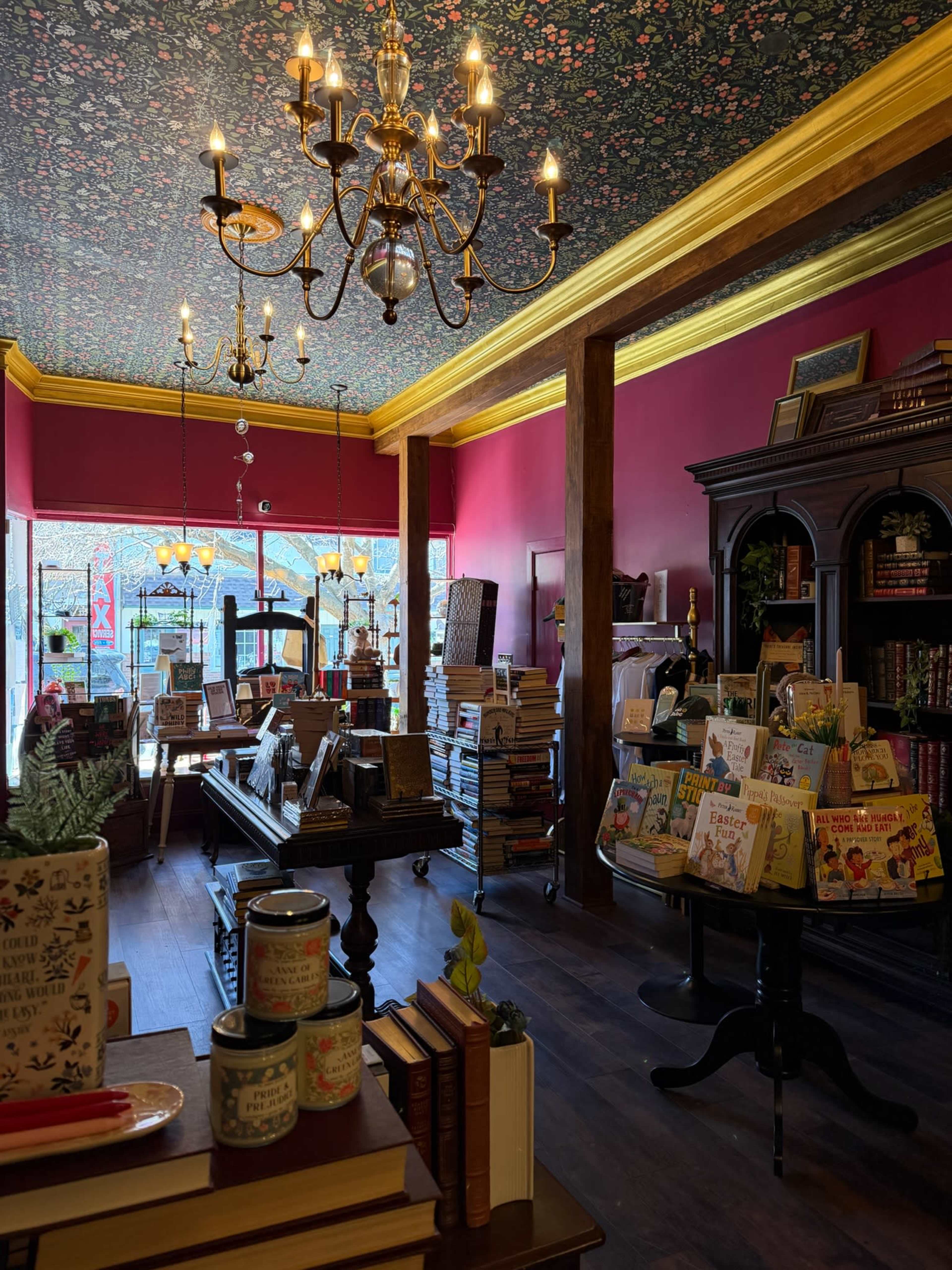 The image shows a cozy bookstore interior with shelves filled with books, a chandelier overhead, and a colorful floral-patterned ceiling.
