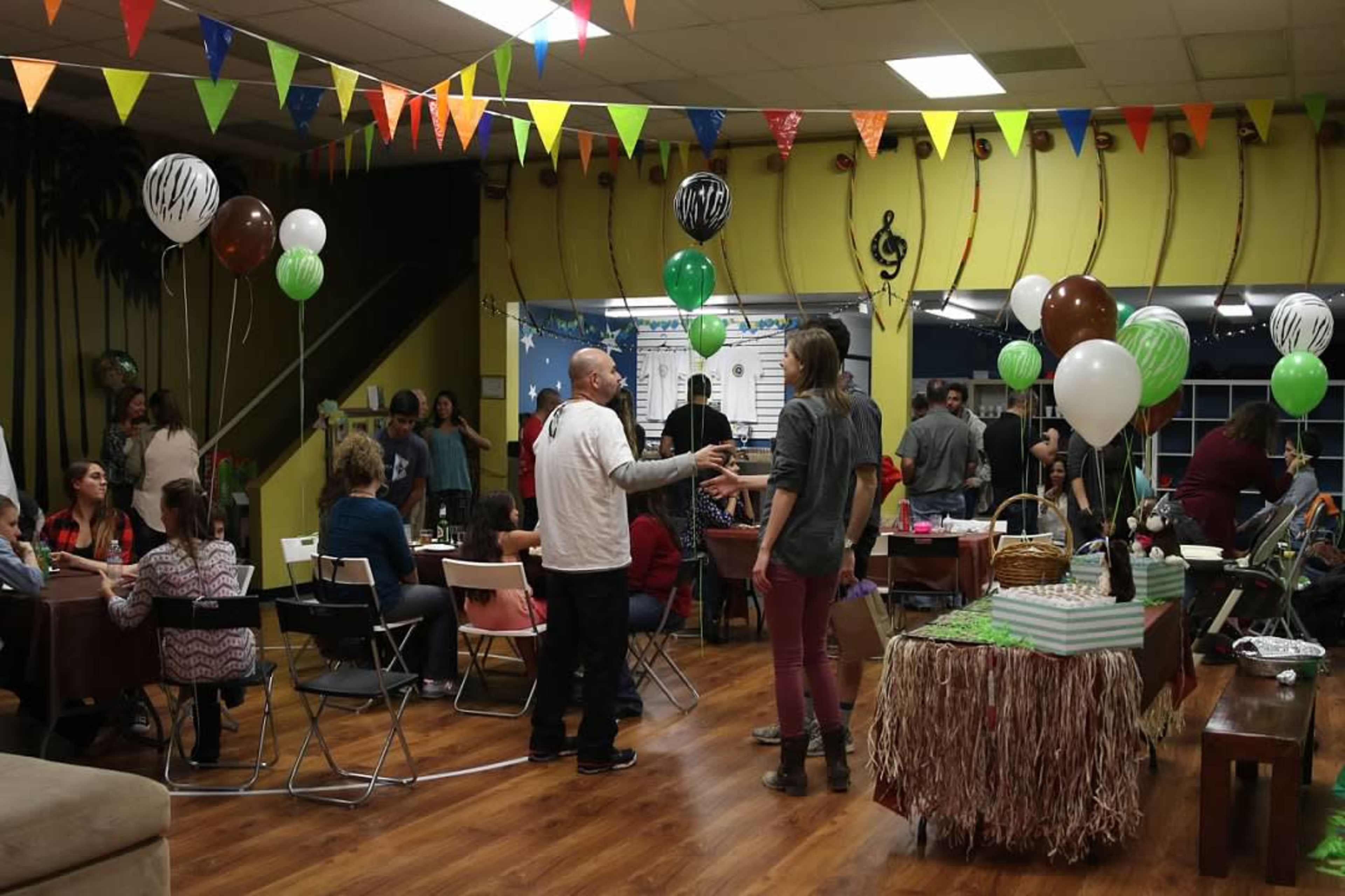 A lively gathering takes place in a decorated room filled with balloons, colorful banners, and people interacting at tables.