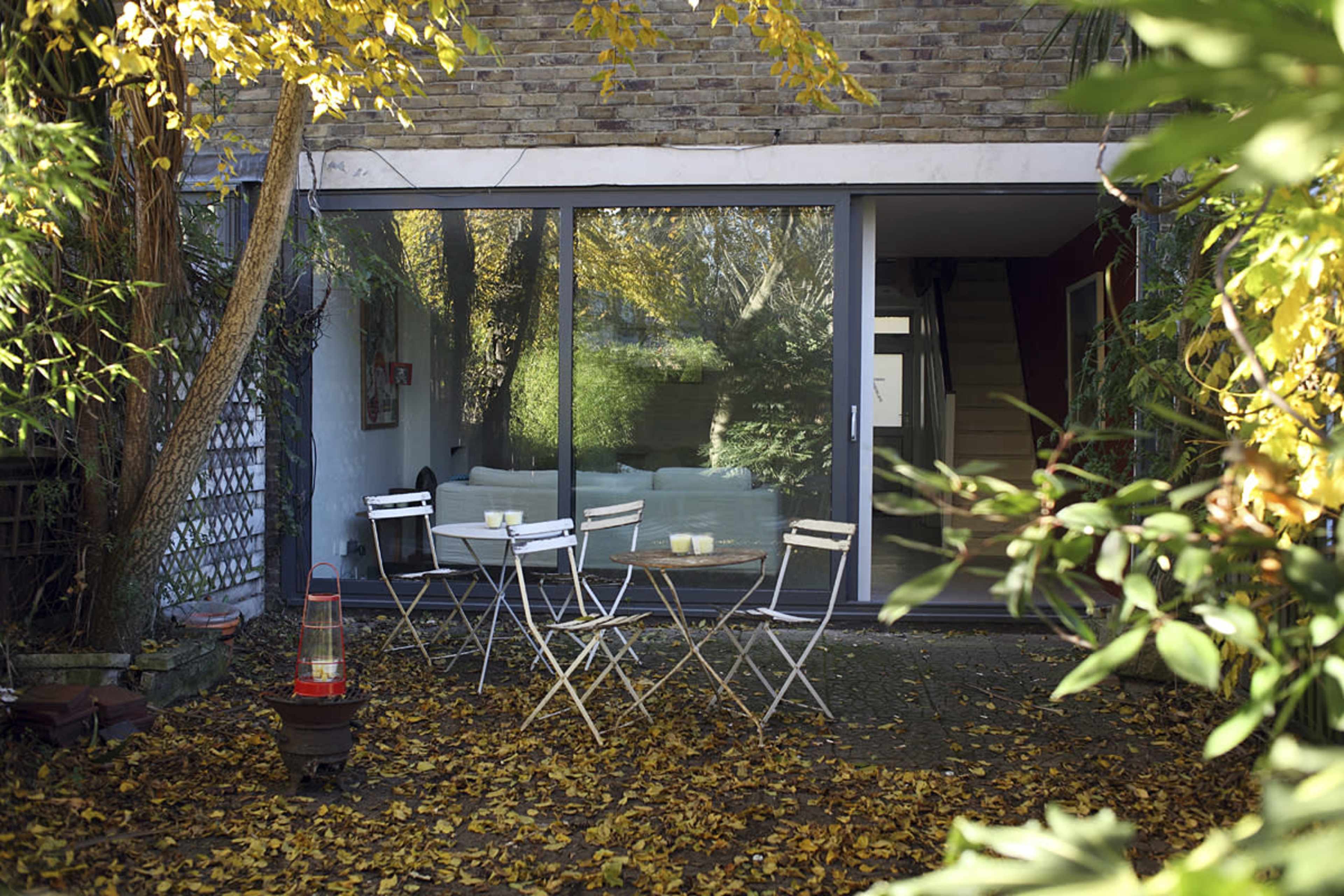 A patio area with six white chairs is surrounded by autumn leaves and tall greenery, leading to a glass sliding door that provides a view into a modern interior space.